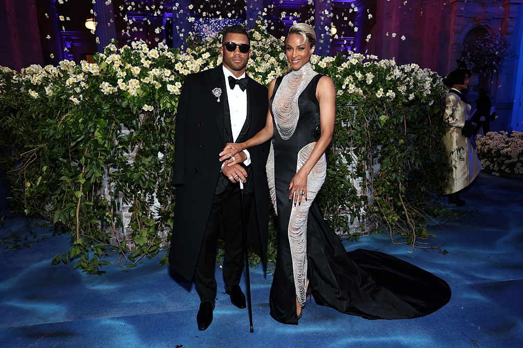 Russell wears a tuxedo with a long coat, and Ciara wears a sleek gown with silver accents