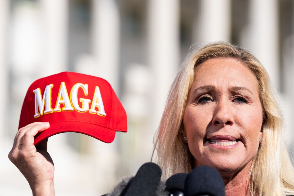 Person holding a hat with "MAGA" text, speaking at a press event