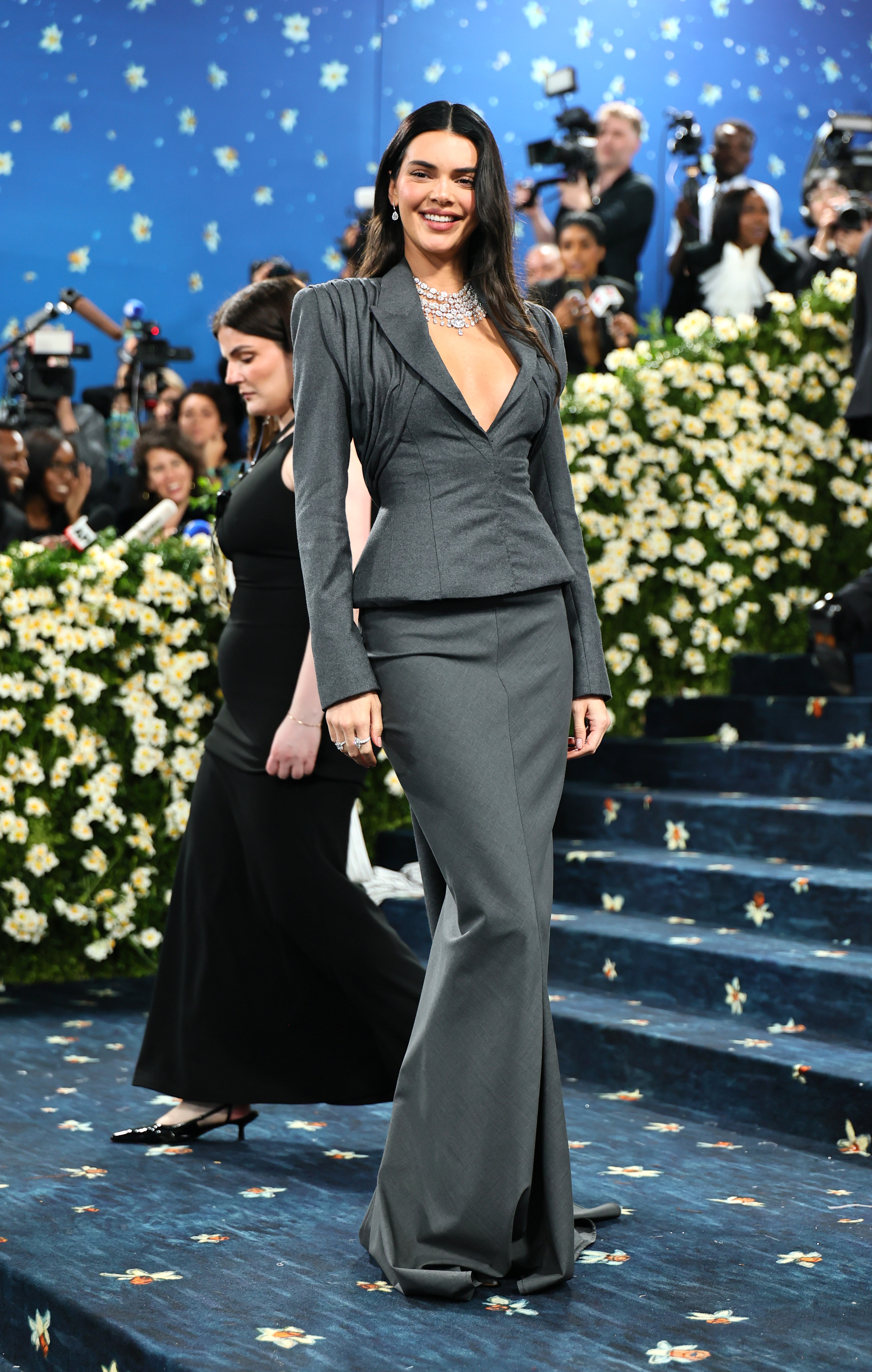 Person in elegant, fitted gown with plunging neckline and statement necklace on a formal event's blue, floral carpet
