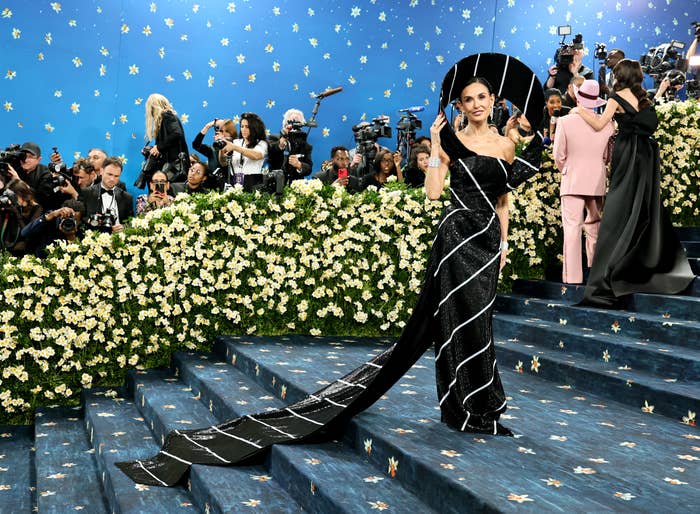 Person in an elegant gown with a long train stands on a flower-decorated staircase at a high-profile event, surrounded by photographers