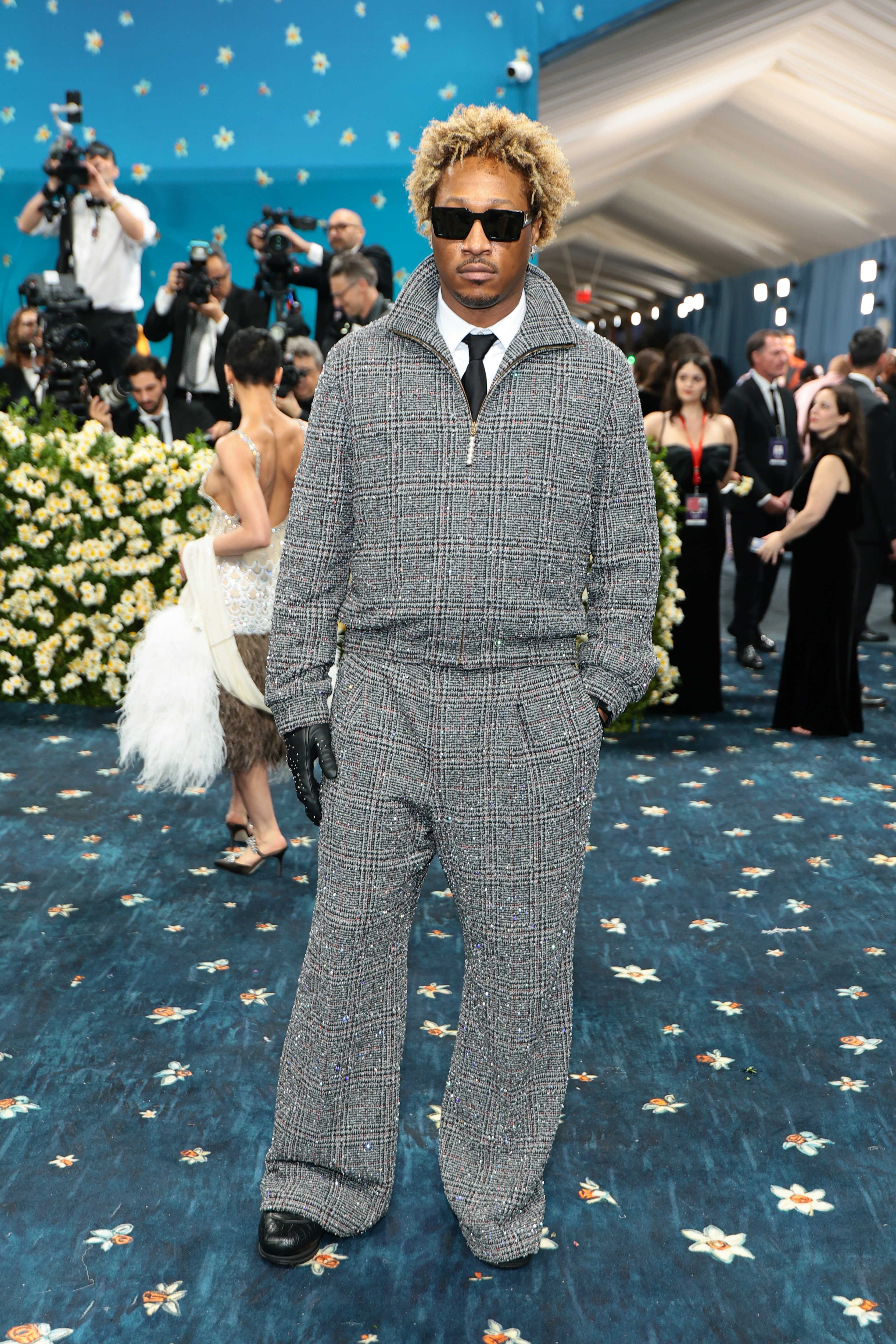 Person wearing a textured suit with wide-legged pants, black gloves, and sunglasses on a glamorous event carpet with floral patterns and photographers