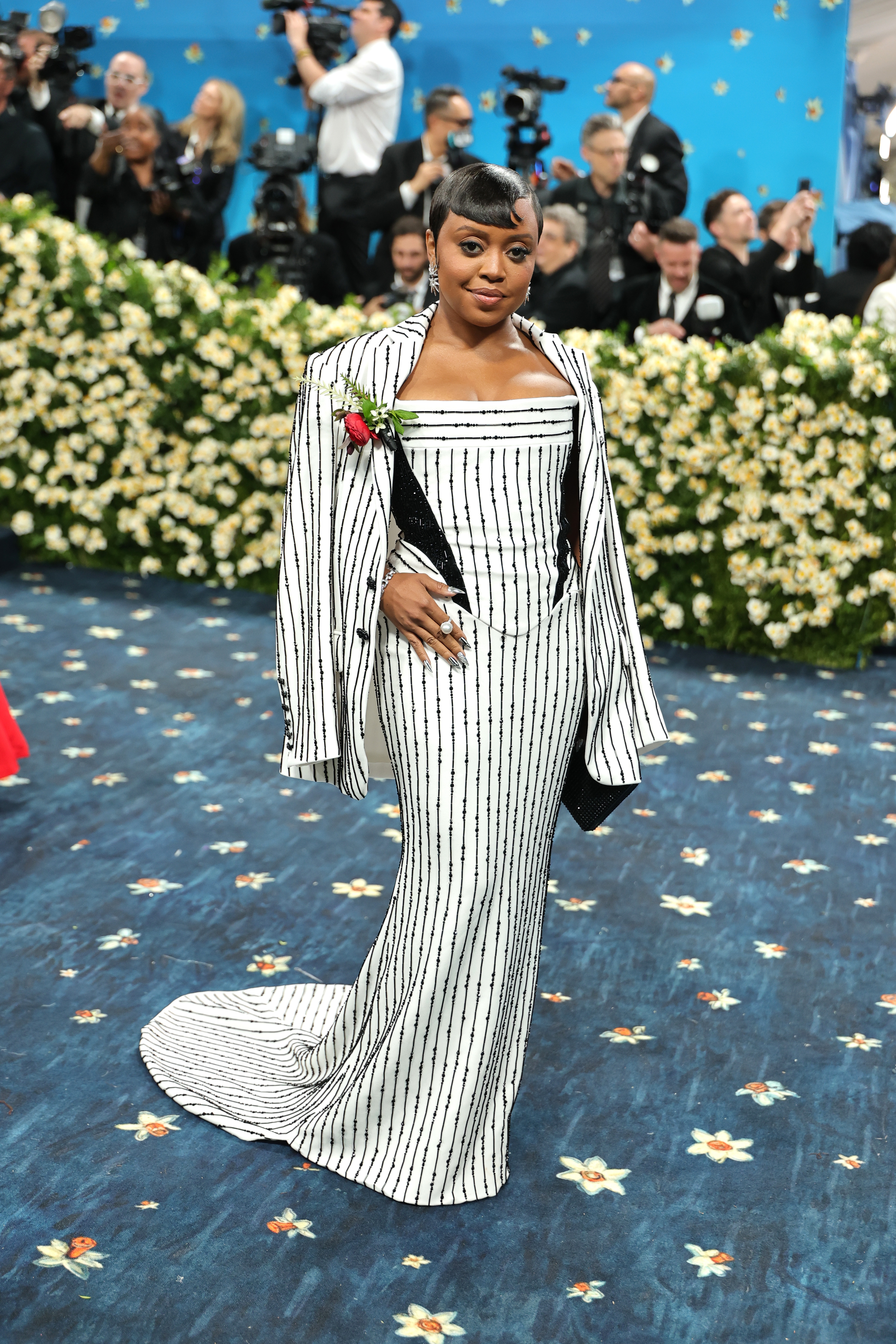 Person in a stylish, black-and-white striped gown with matching cape, holding clutch on floral-patterned carpet at a celebrity event