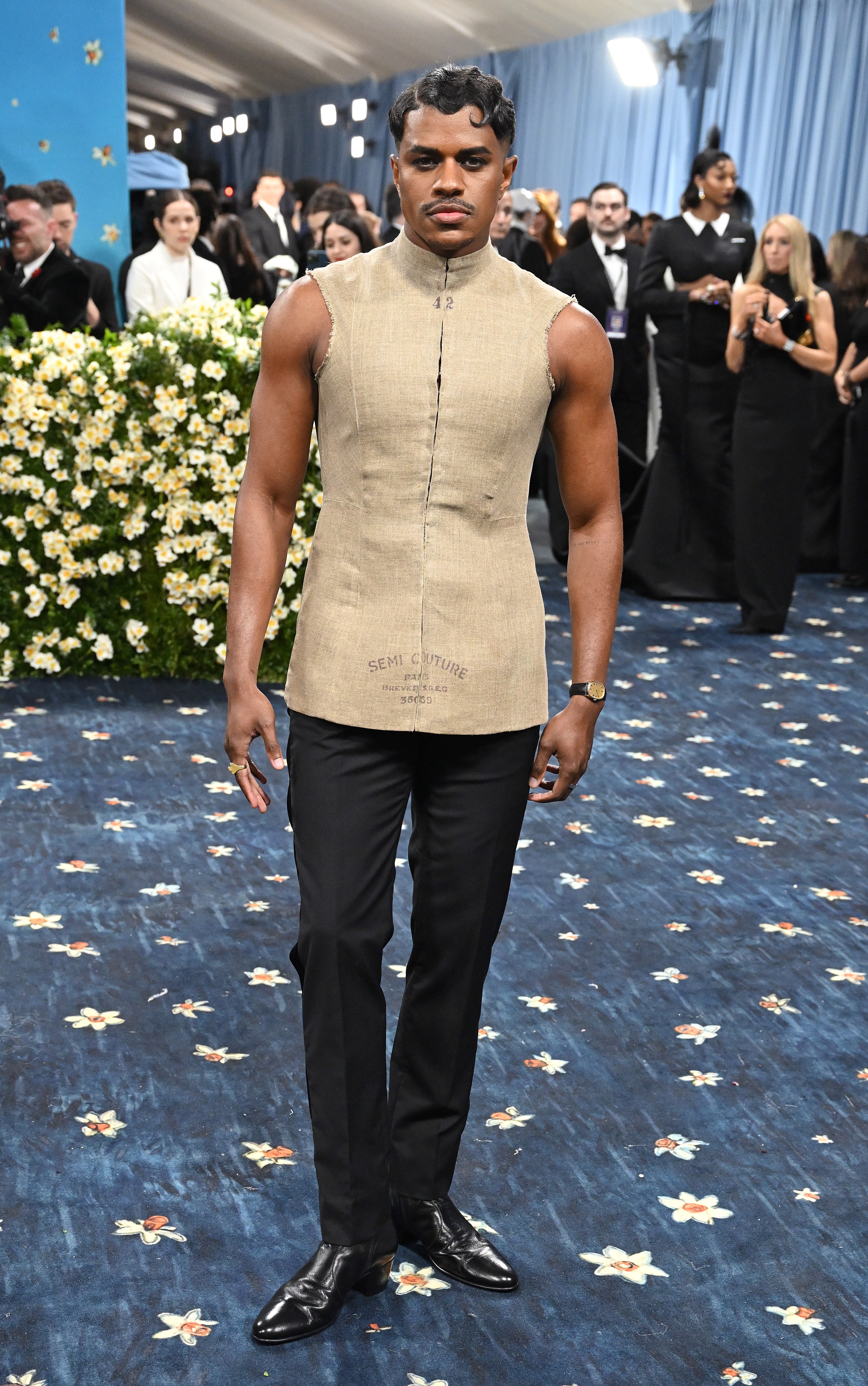 A person in a sleeveless, fitted top and black pants poses on a formal event's floral-patterned carpet, surrounded by attendees
