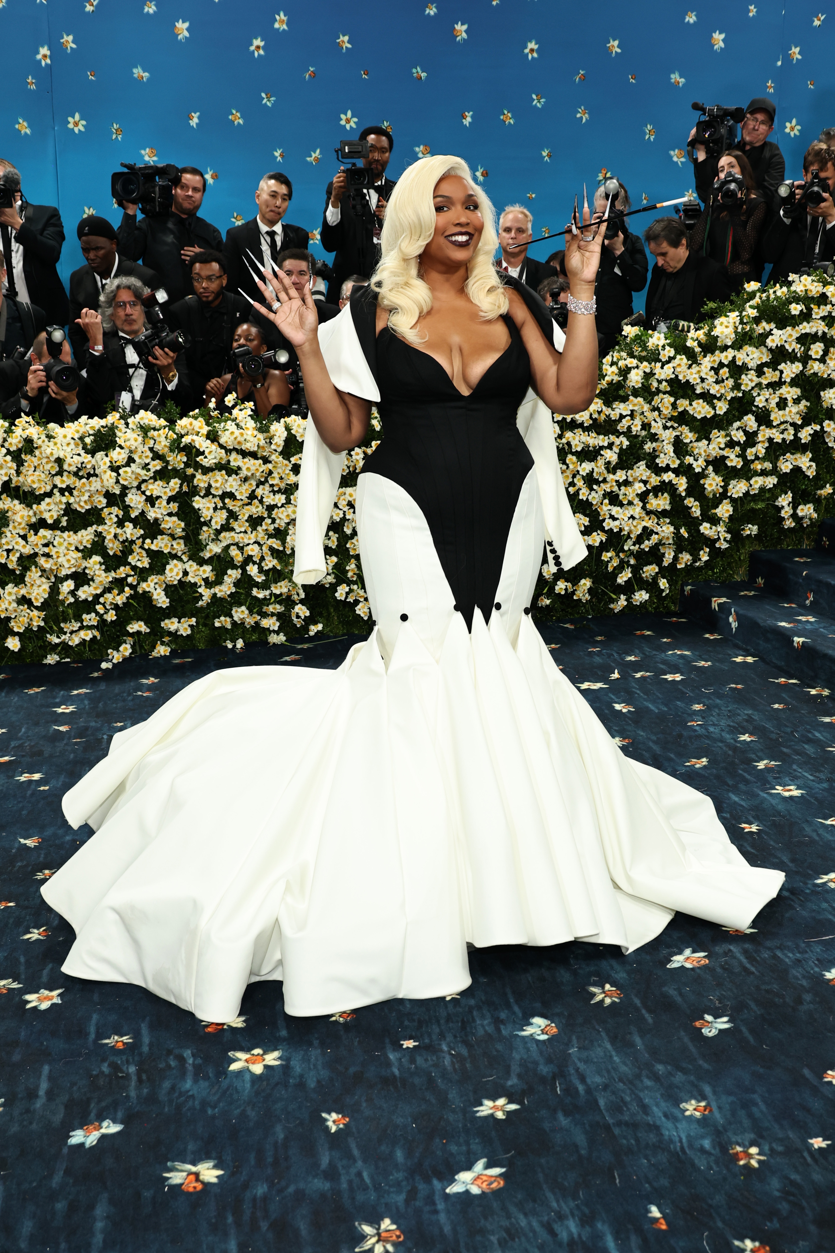 Person in a dramatic black and white gown posing on a floral-lined event carpet, smiling with photographers capturing the moment