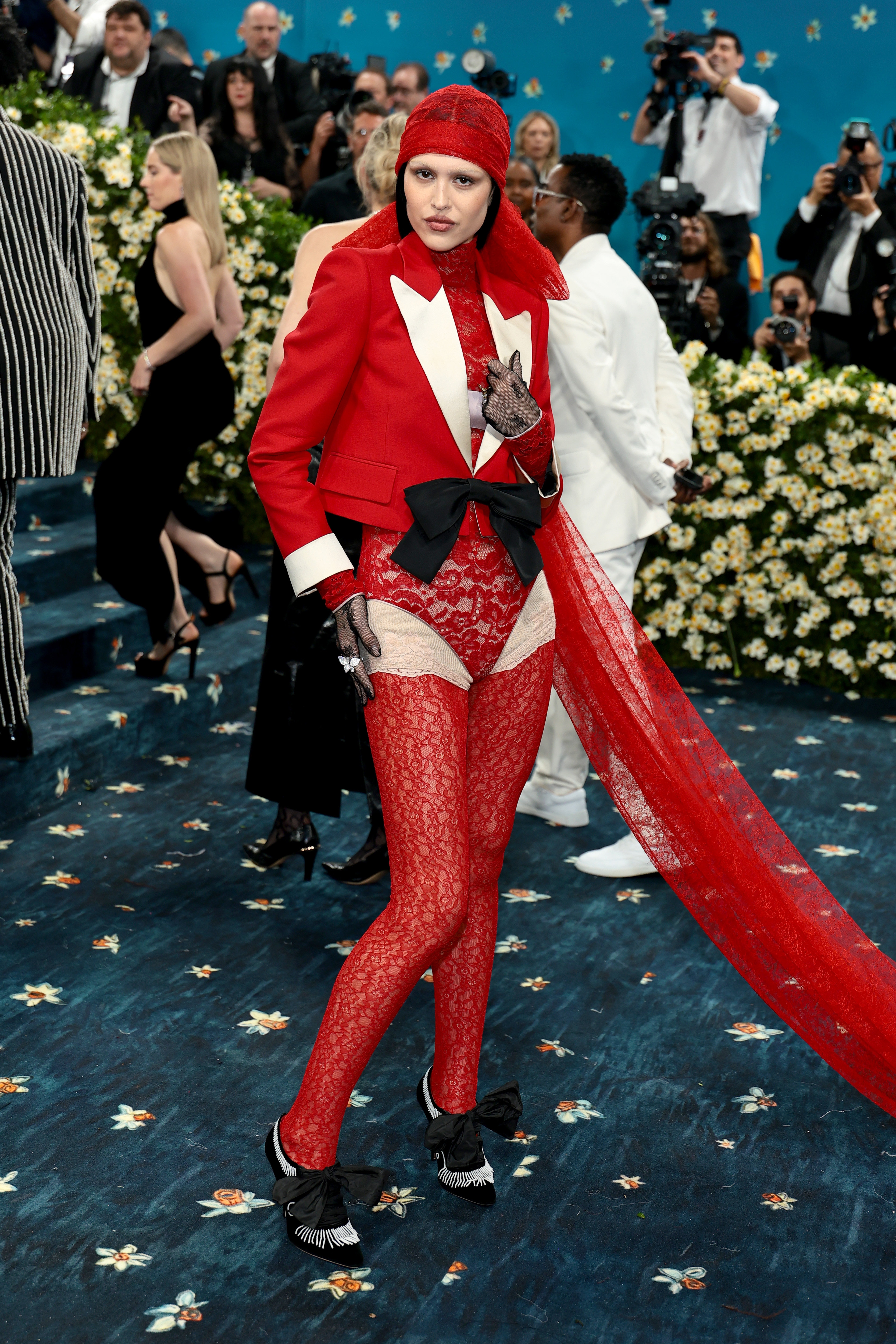 Person in a red lace ensemble with a matching veil and bow, posing on a floral-themed event carpet while photographers capture the scene