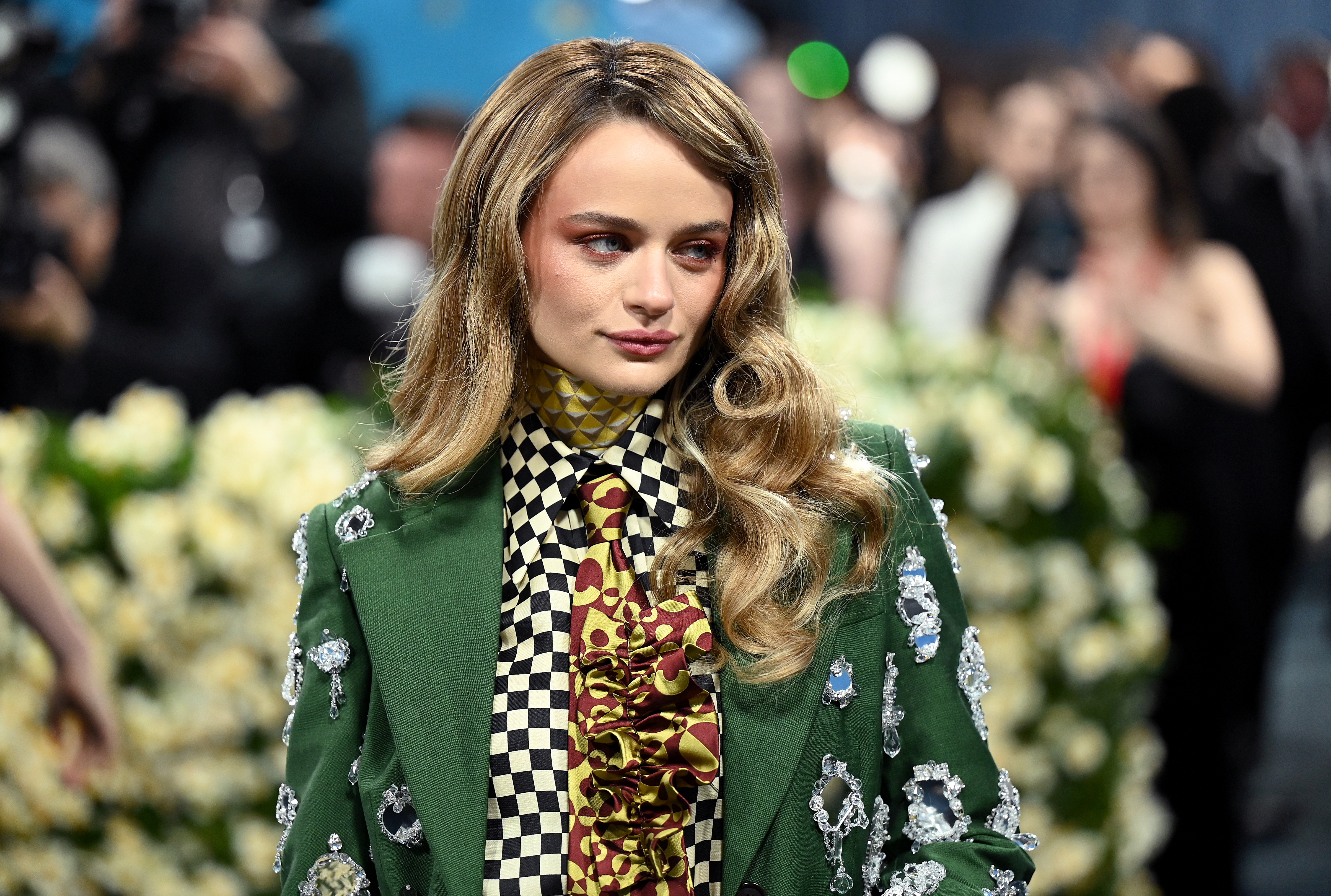 Person in patterned outfit with long hair, posing at a formal event. Background features flowers and blurred attendees