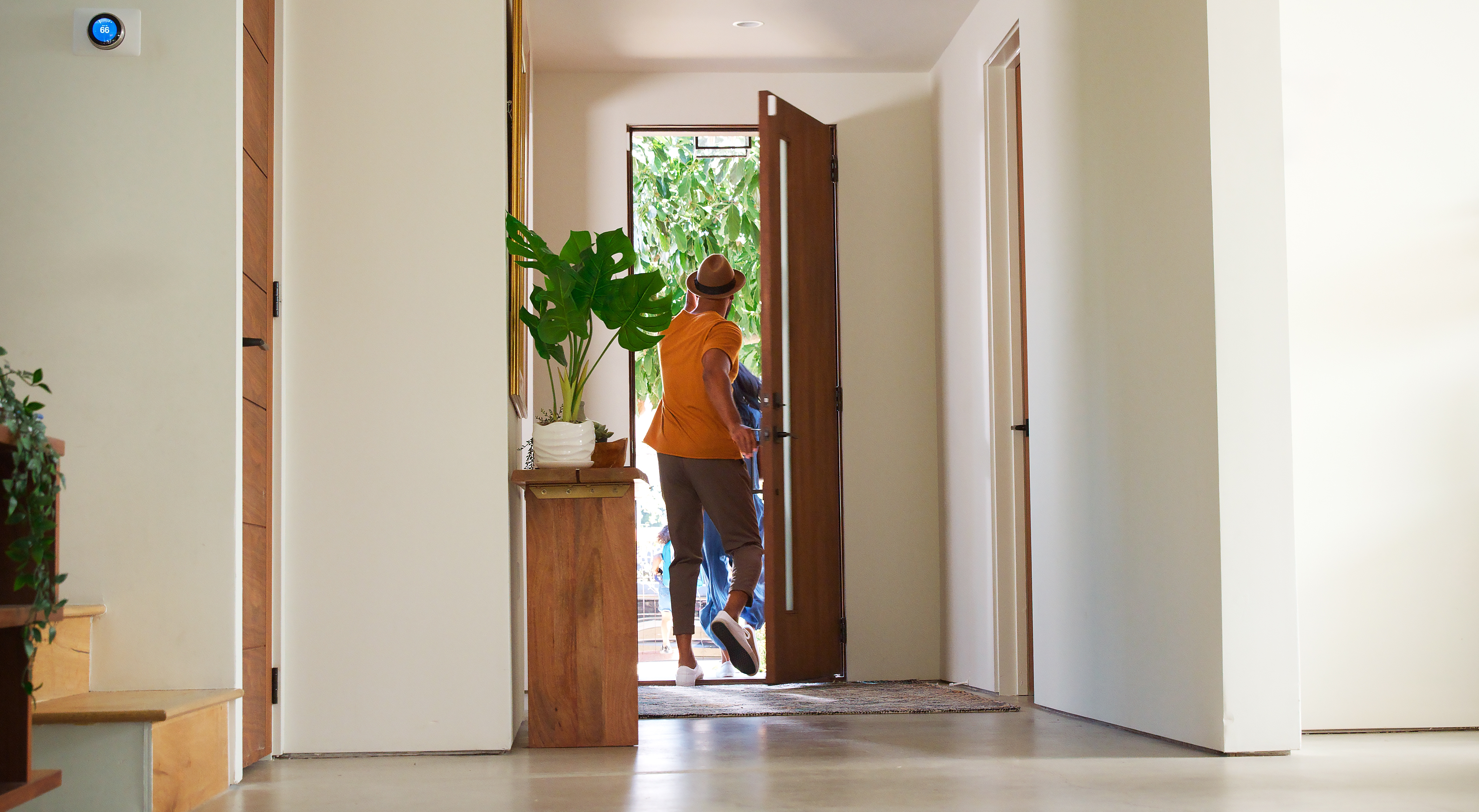 Person in casual outfit and hat leaving a bright, modern home through an open door, with plants by the entryway