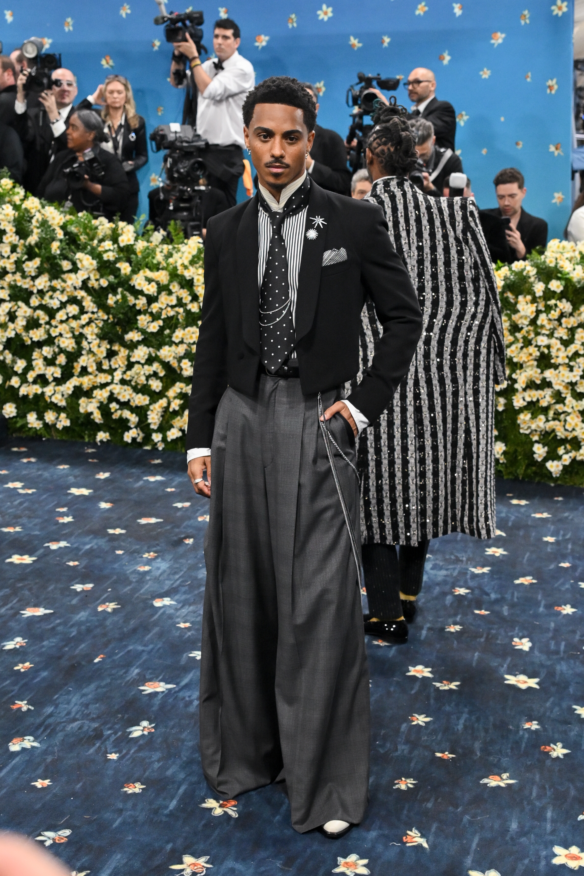 Person in a stylish black and white ensemble with a tailored jacket and wide-leg trousers at a celebrity event. Photographers in the background