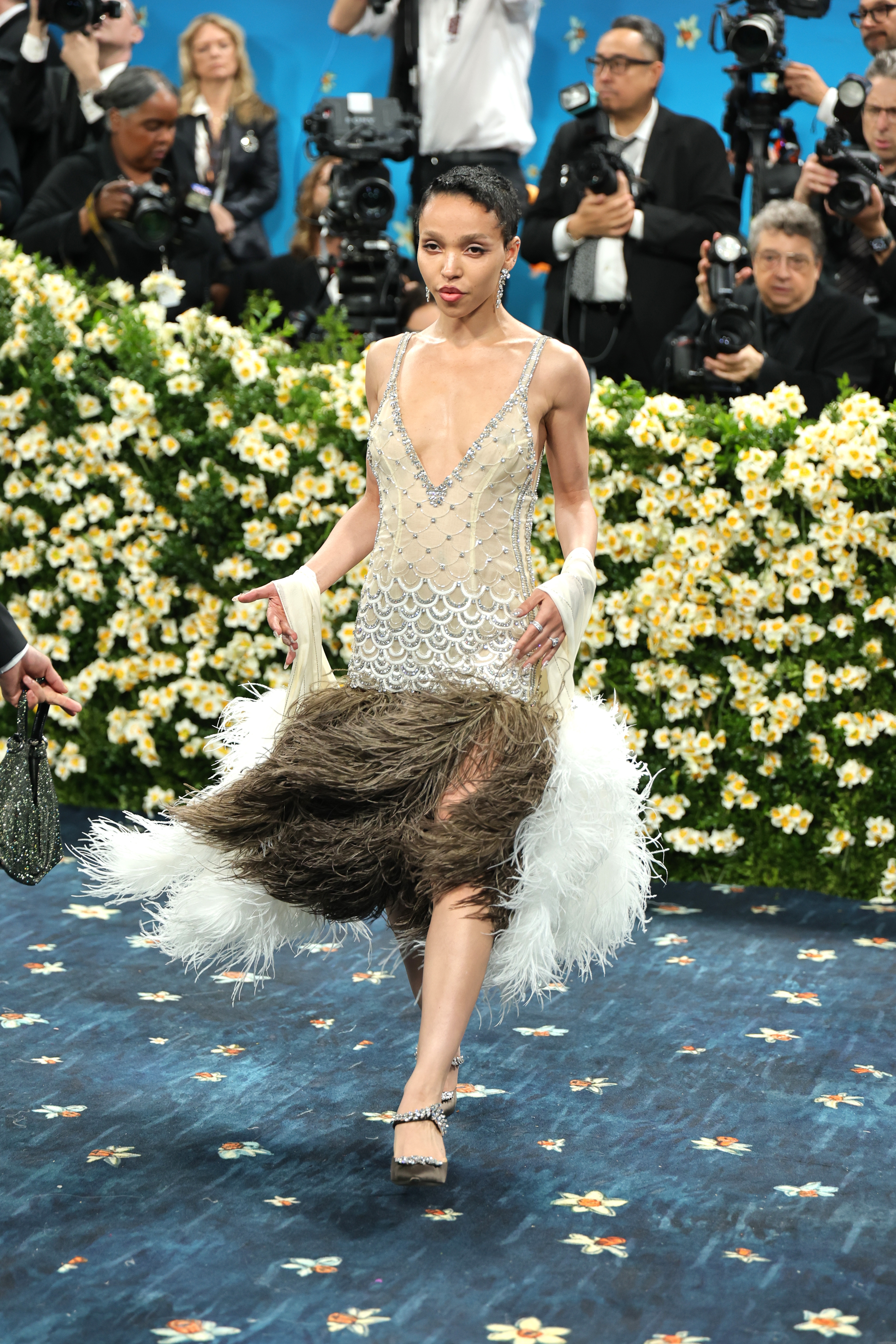 A person on a floral-themed red carpet wearing a sparkling dress with feathered details, surrounded by photographers