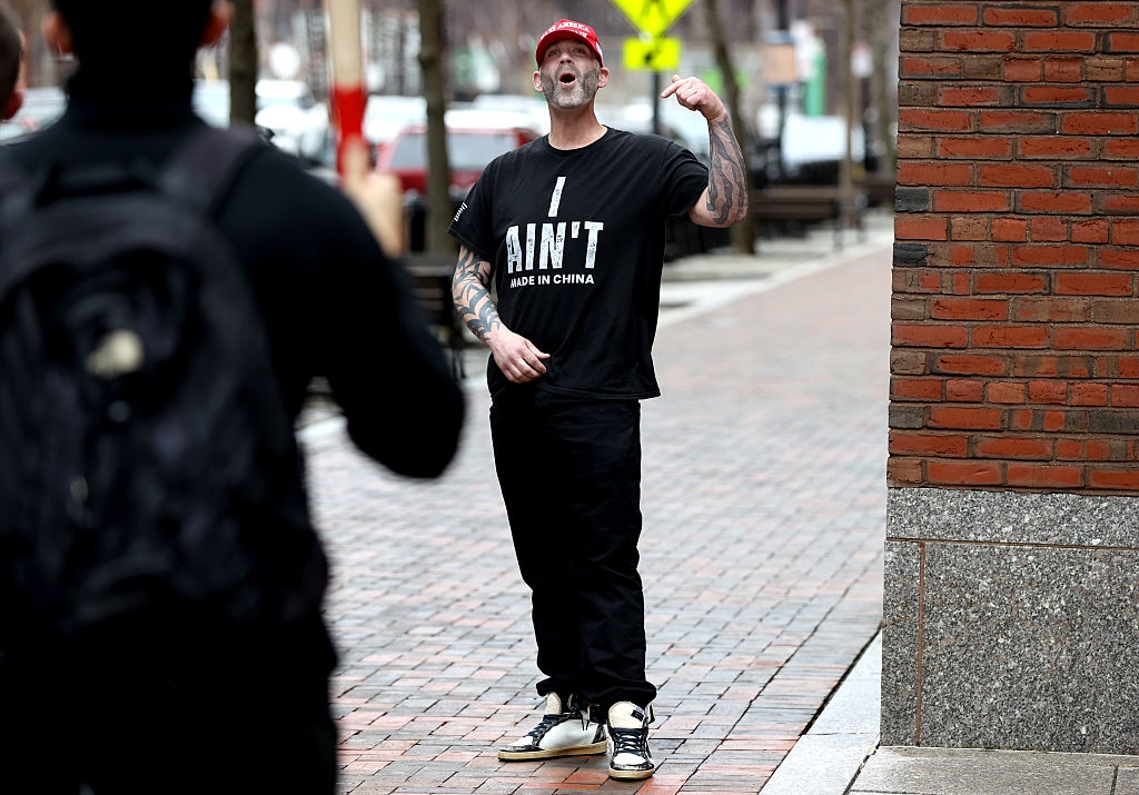 A person gestures energetically on a street, wearing a cap and a T-shirt with the text "I Ain't Made in China."