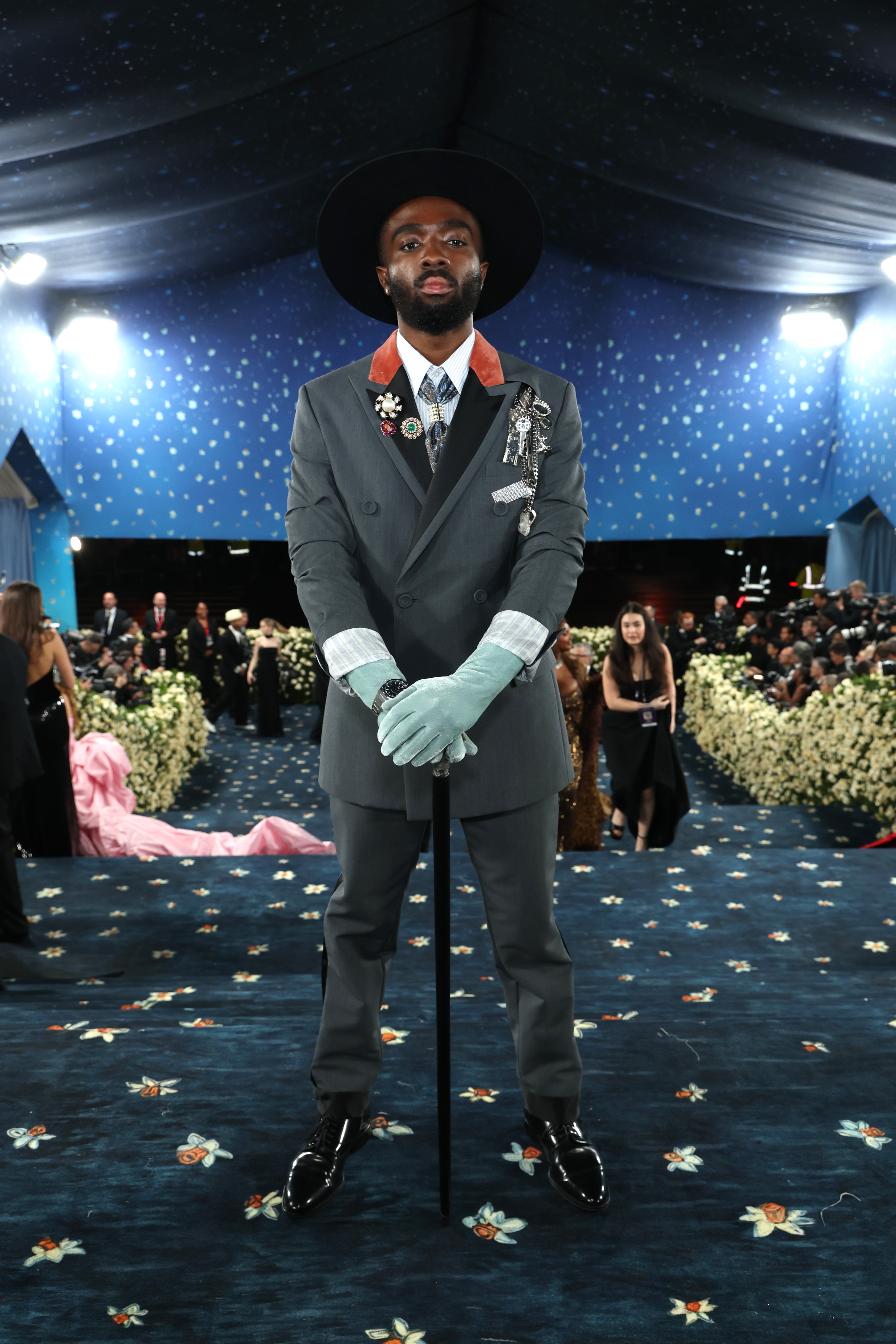 Person in a stylish gray suit with medals, pale gloves, and a wide-brim hat stands confidently on a floral-patterned carpet at a formal event