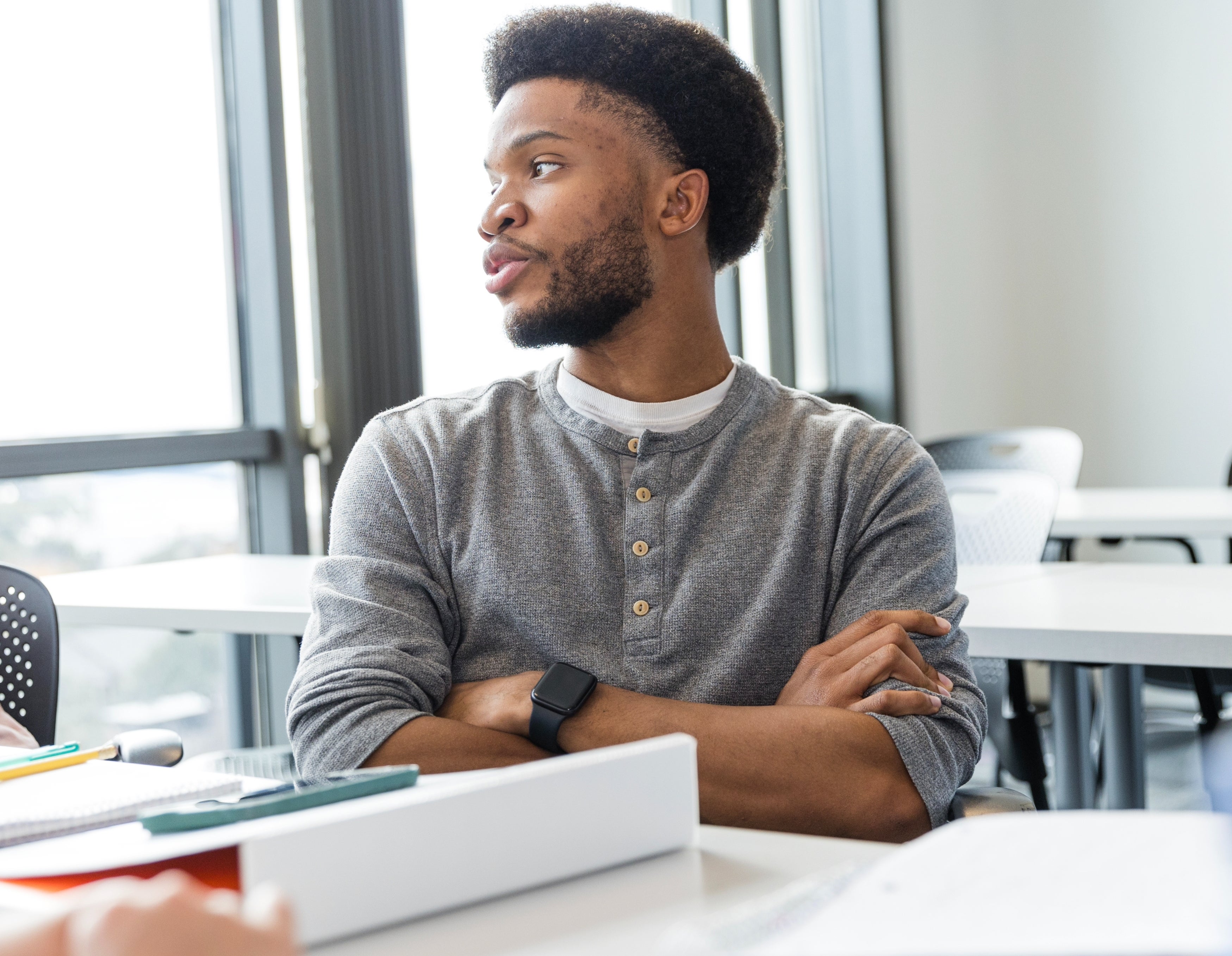 A man in a gray sweater sits at a table in a meeting room, looking to the side, with people seated around him