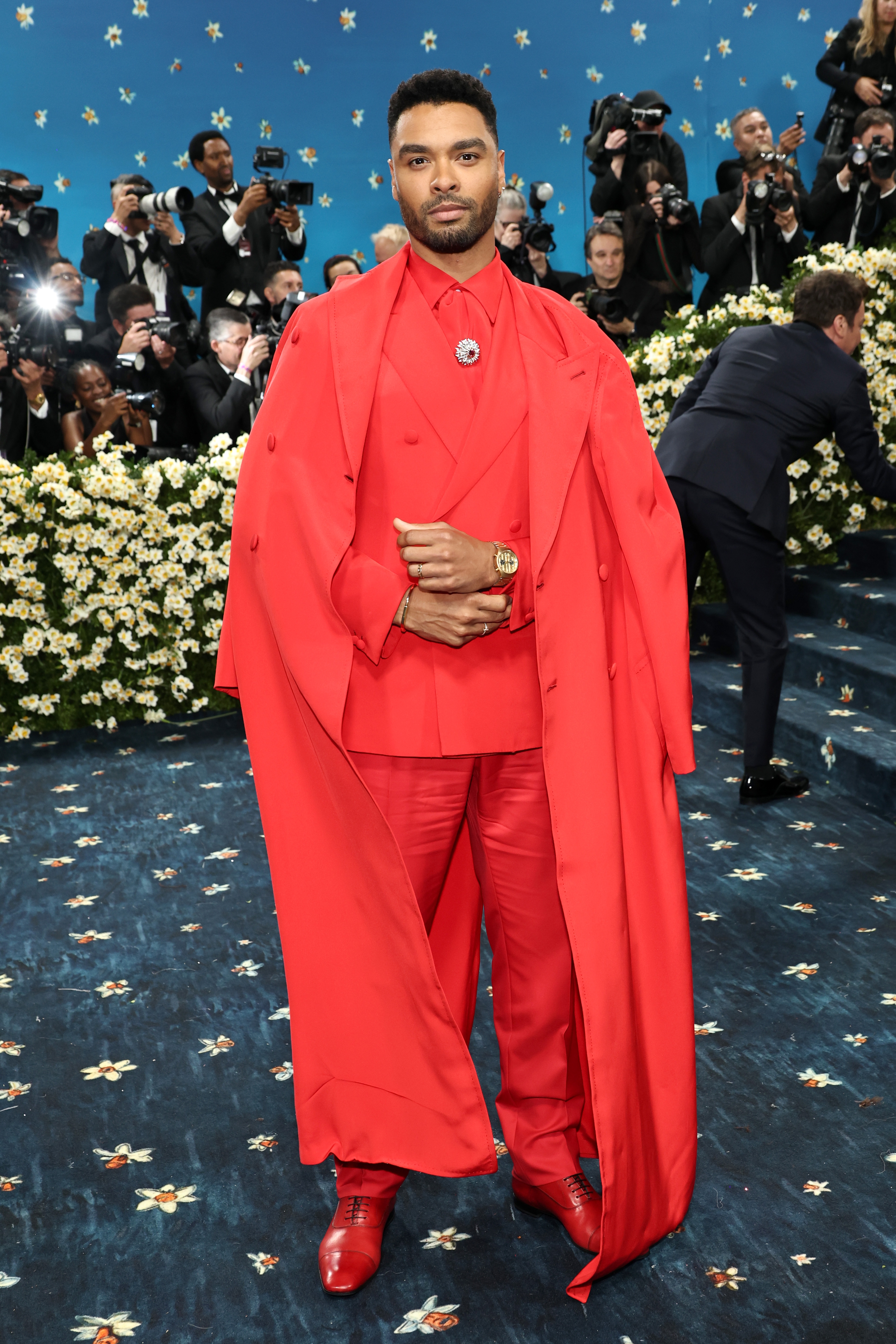 Person wearing a layered, tailored red ensemble with a long coat and matching shoes on a floral-lined event carpet, surrounded by photographers