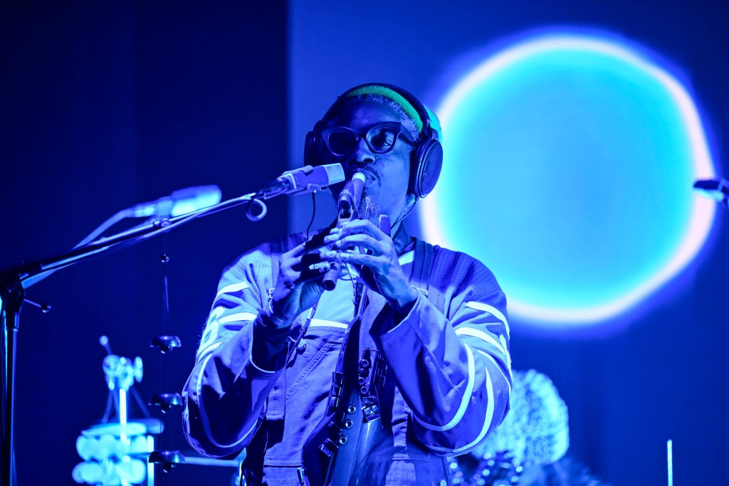 Musician performs on stage holding a woodwind instrument, wearing a jacket and headphones. A bright circular light is in the background