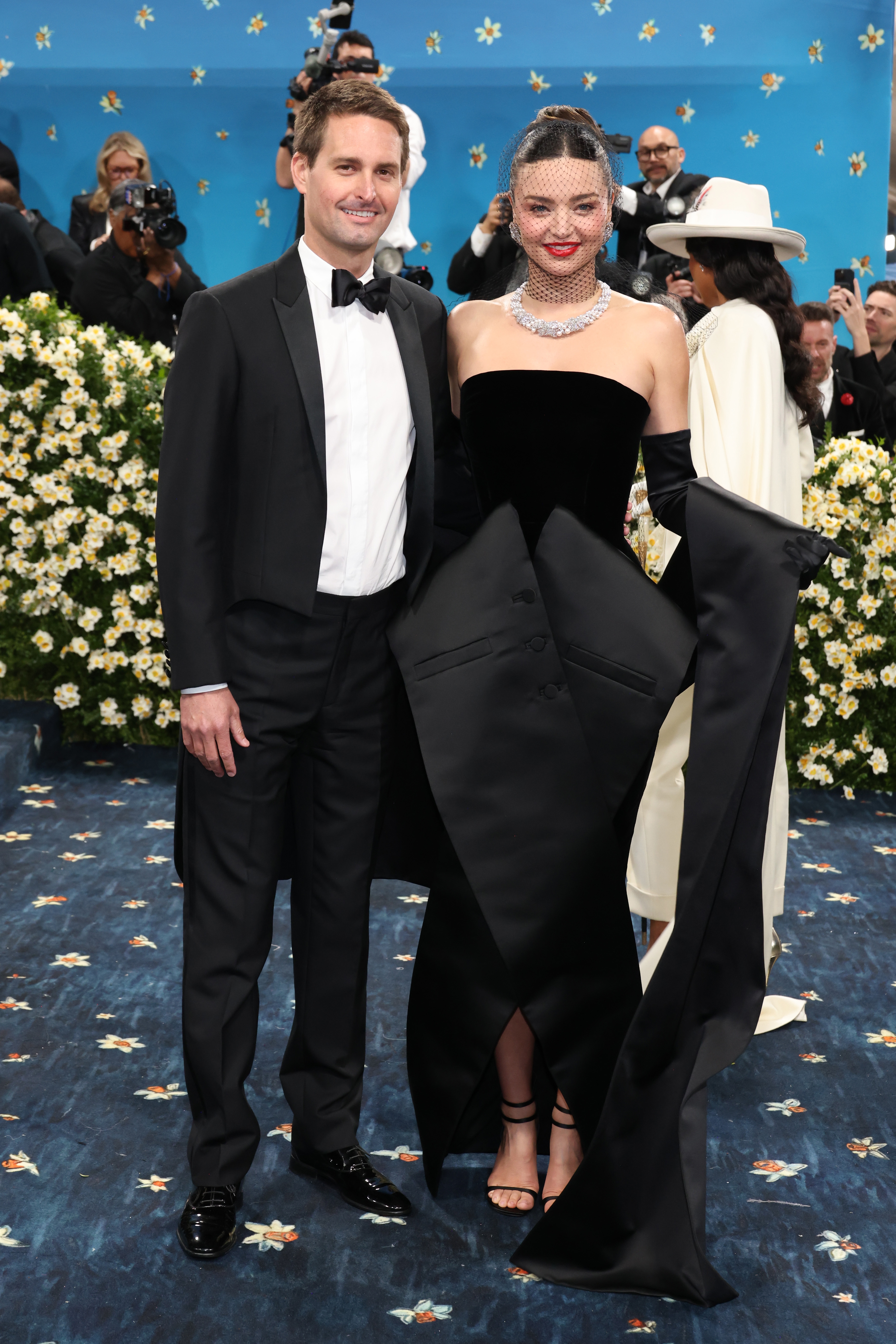 Two people pose on the red carpet; one in a tuxedo and the other in a black velvet gown with satin gloves, surrounded by photographers and floral decor