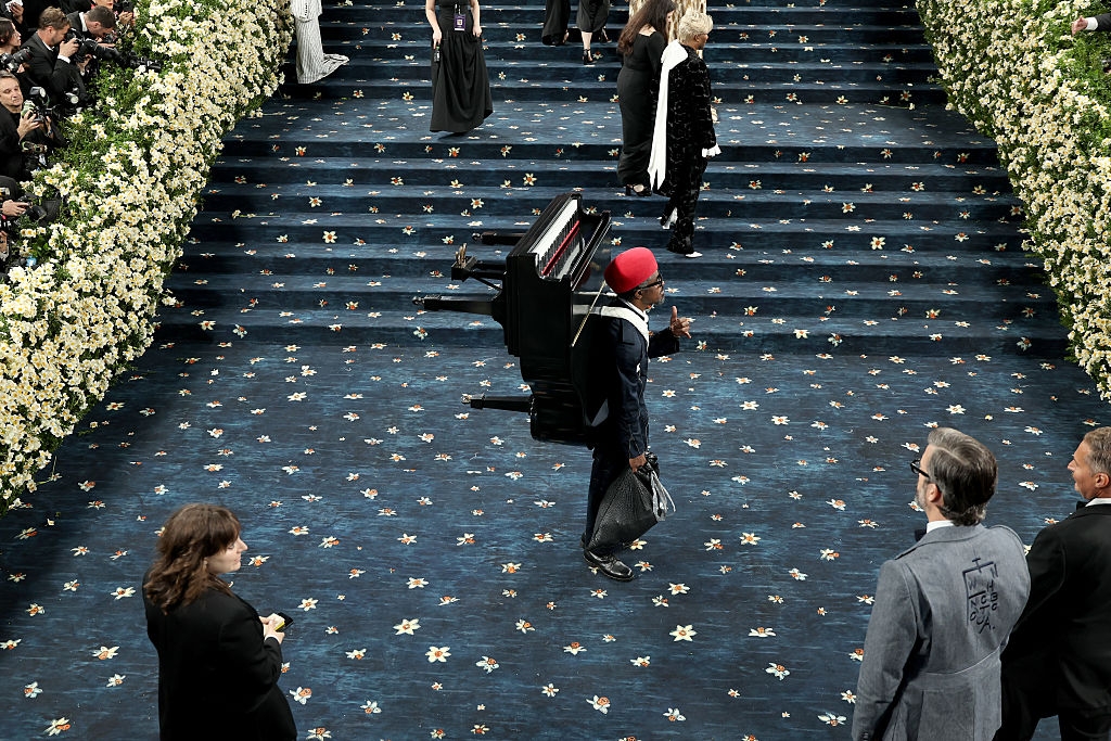 Man carrying a piano on his back at a formal event with floral decorations and people in elegant attire