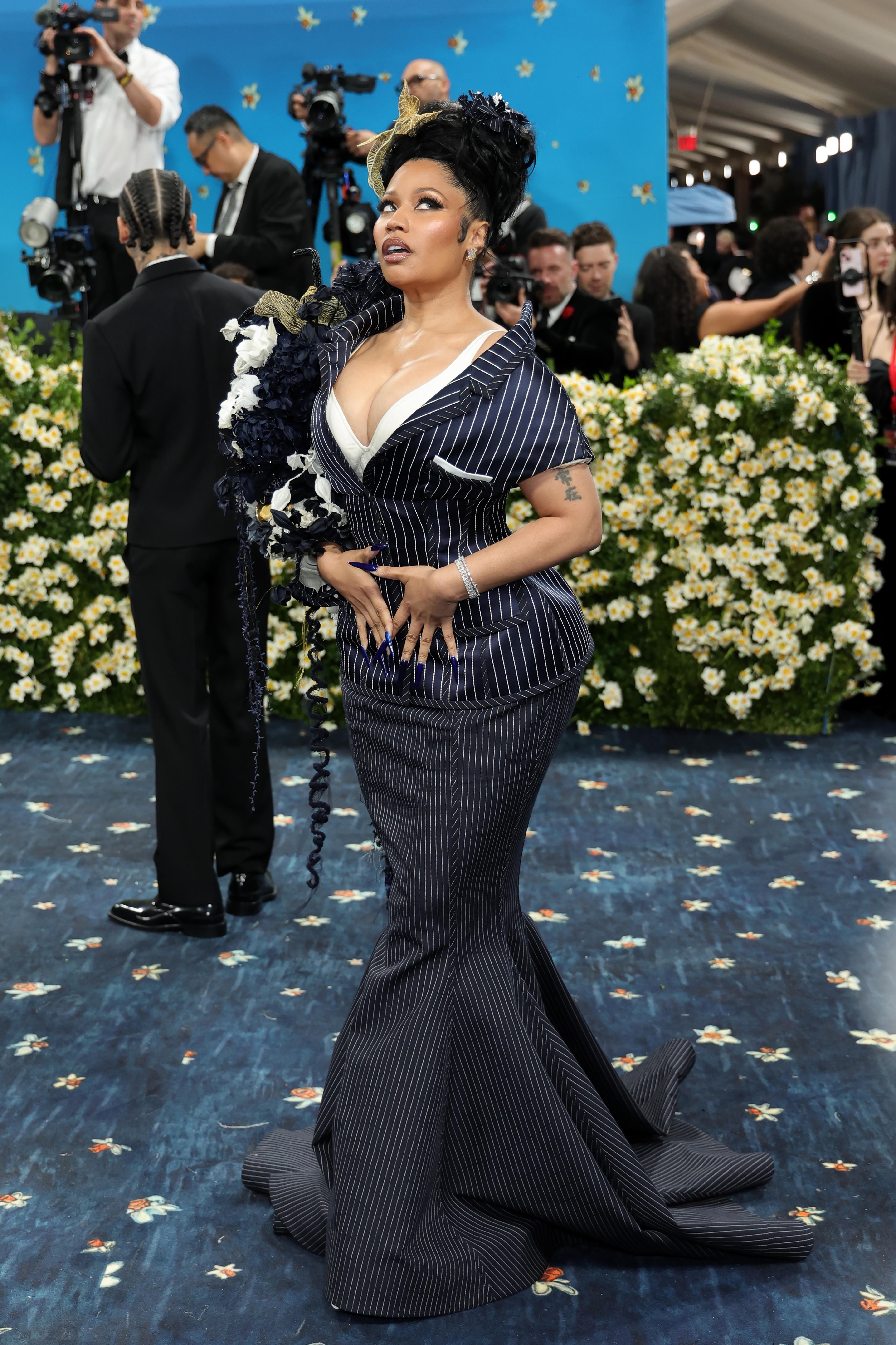 Person on a formal blue carpet in a pinstriped gown with floral details, posing in front of photographers and floral backdrop