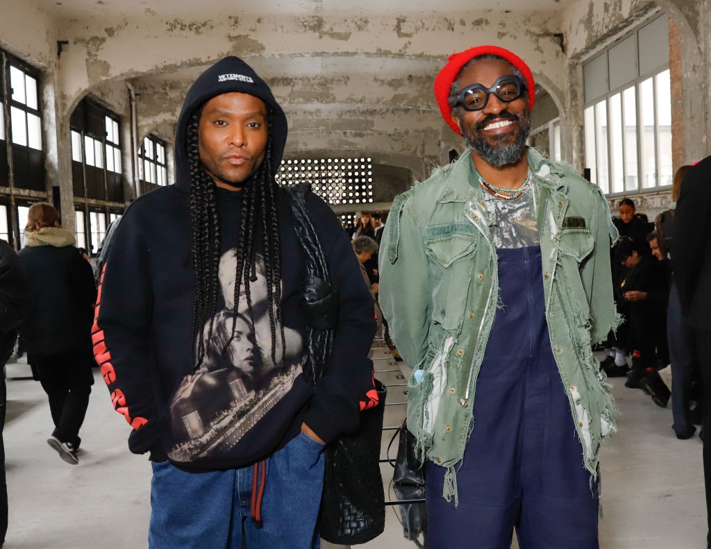 Two people in casual, artistic style clothing at an indoor event, one wearing a graphic hoodie and the other in a distressed jacket with a red hat