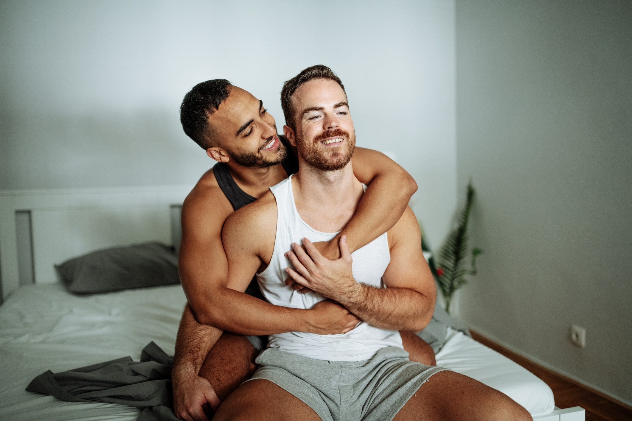 Two people sit closely on a bed, smiling and embracing each other affectionately, conveying a sense of intimacy and connection