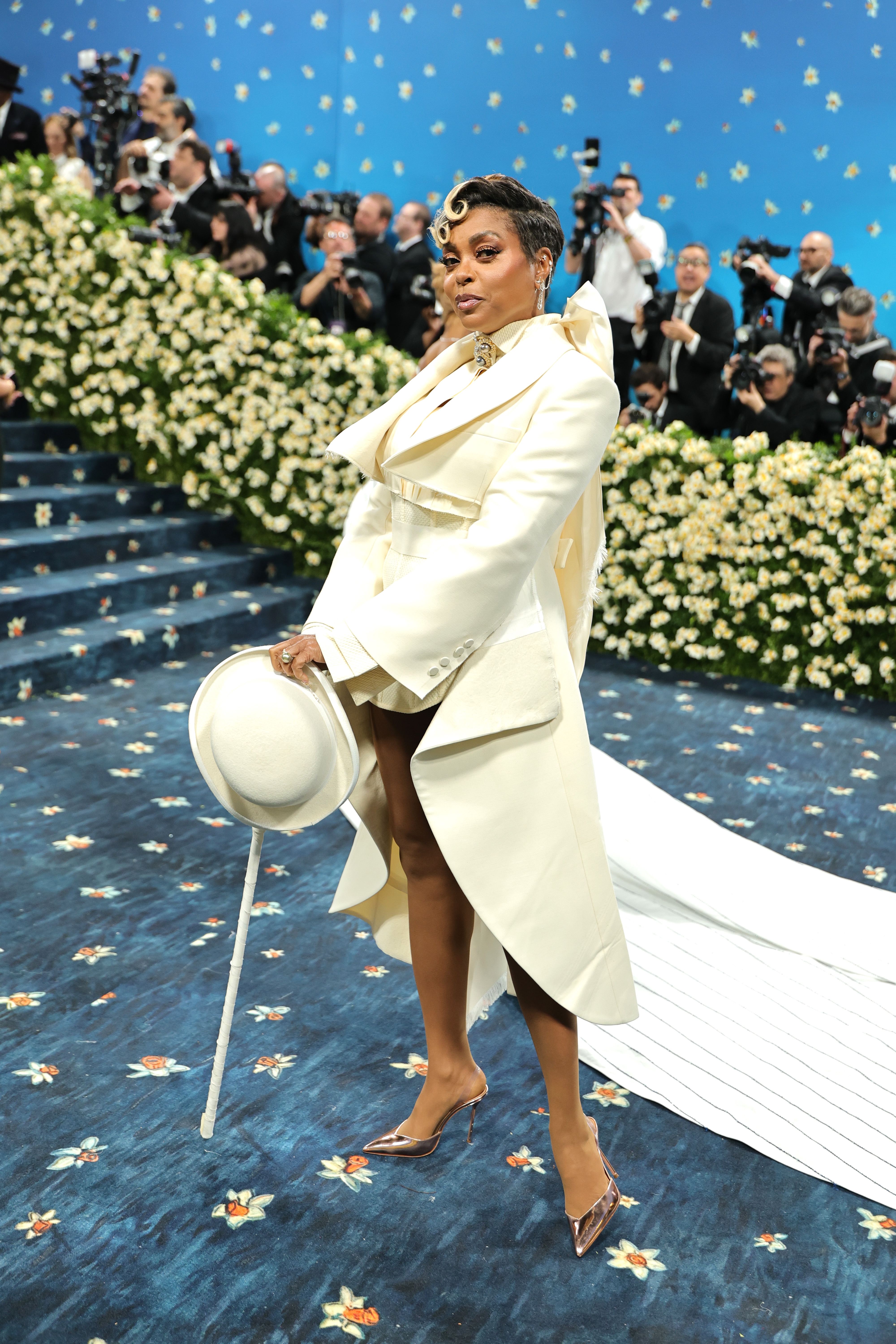 A person on a floral-patterned carpet wearing an elegant white suit with an asymmetrical train, holding a wide-brimmed hat, surrounded by photographers
