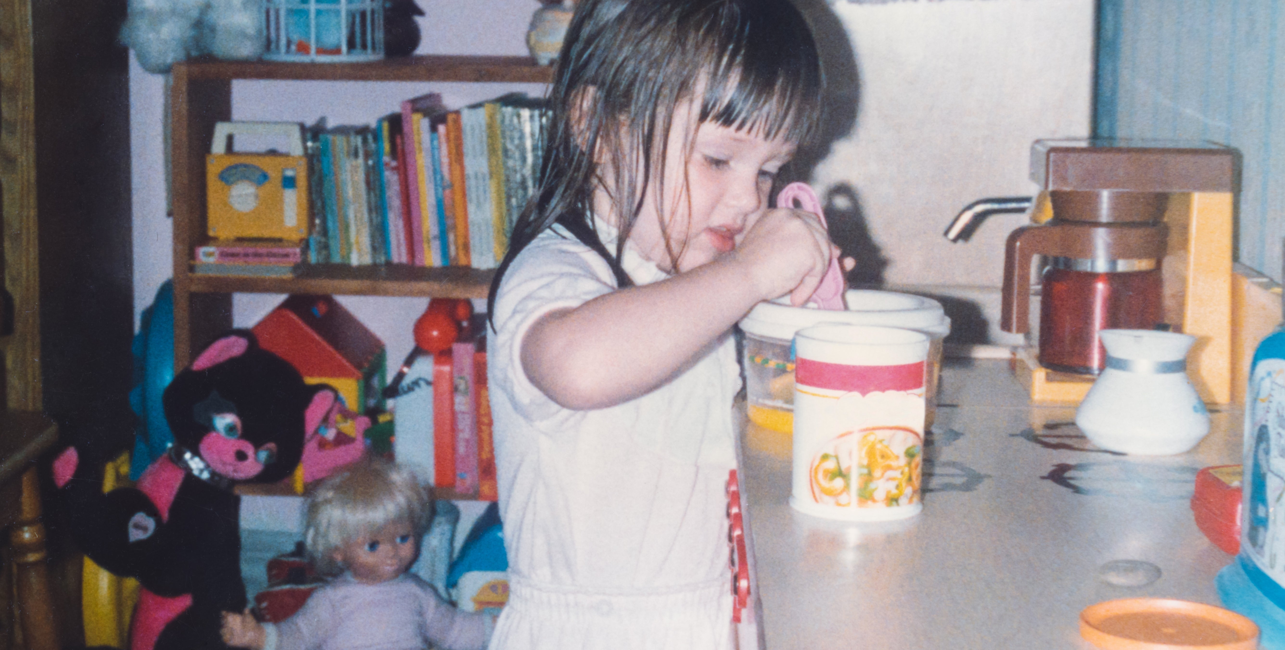 A young child plays in a toy kitchen, surrounded by stuffed animals and books, focused on her activity
