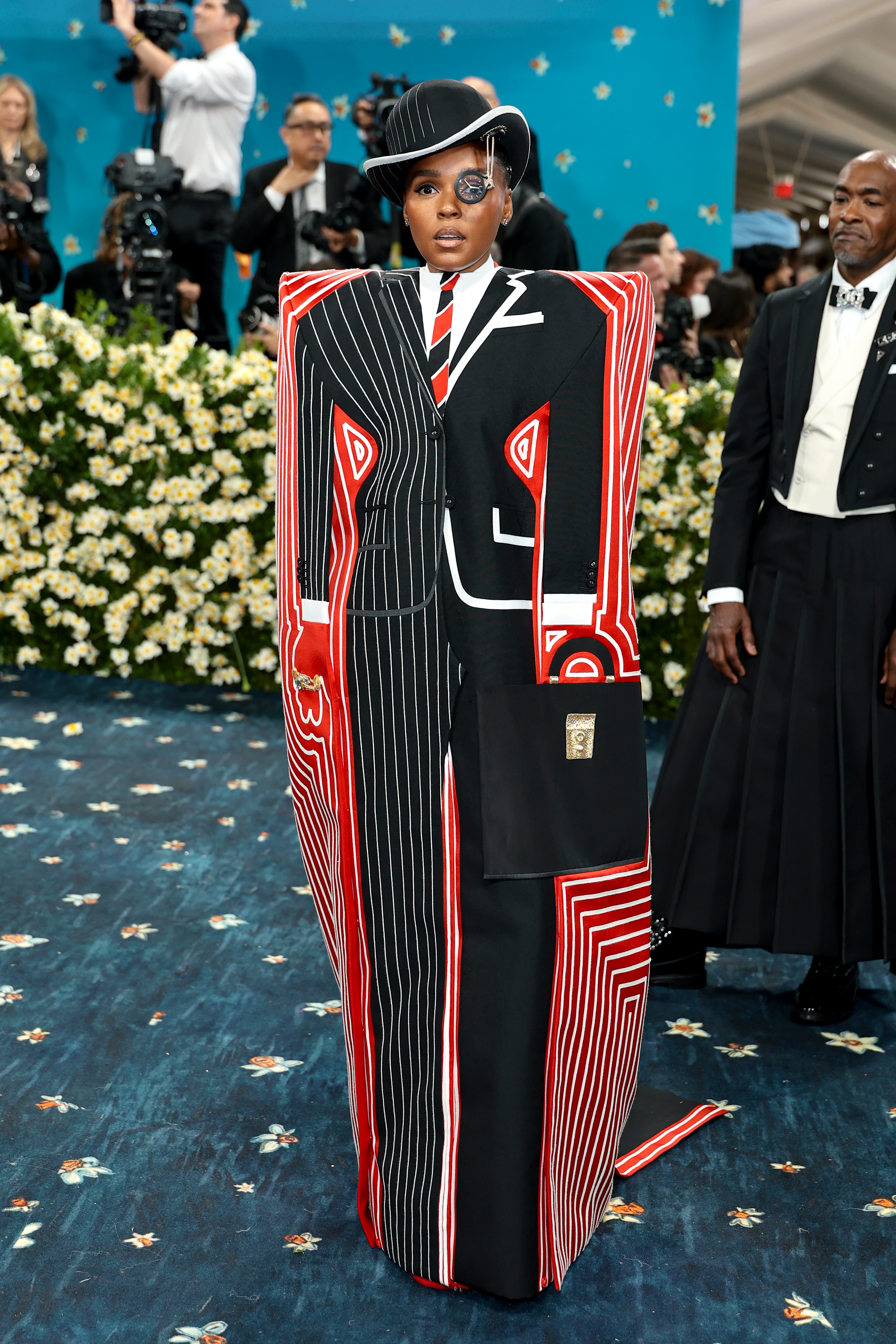 Person on a blue carpet wearing a bold, geometric suit with a hat, surrounded by photographers and attendees in formal wear