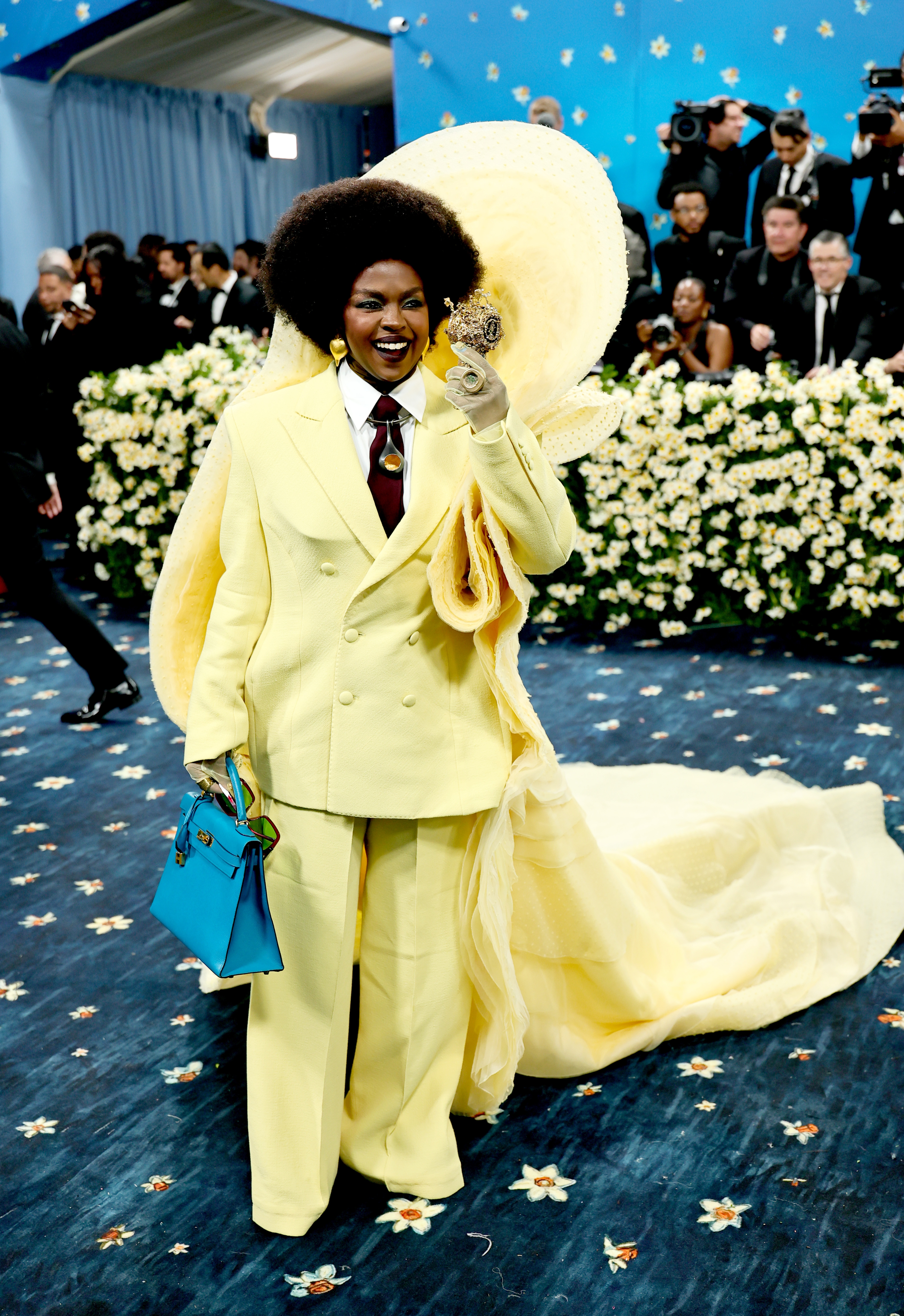 Person in a flowing outfit with a large circular headpiece poses on red carpet holding up a ring, carrying a blue handbag