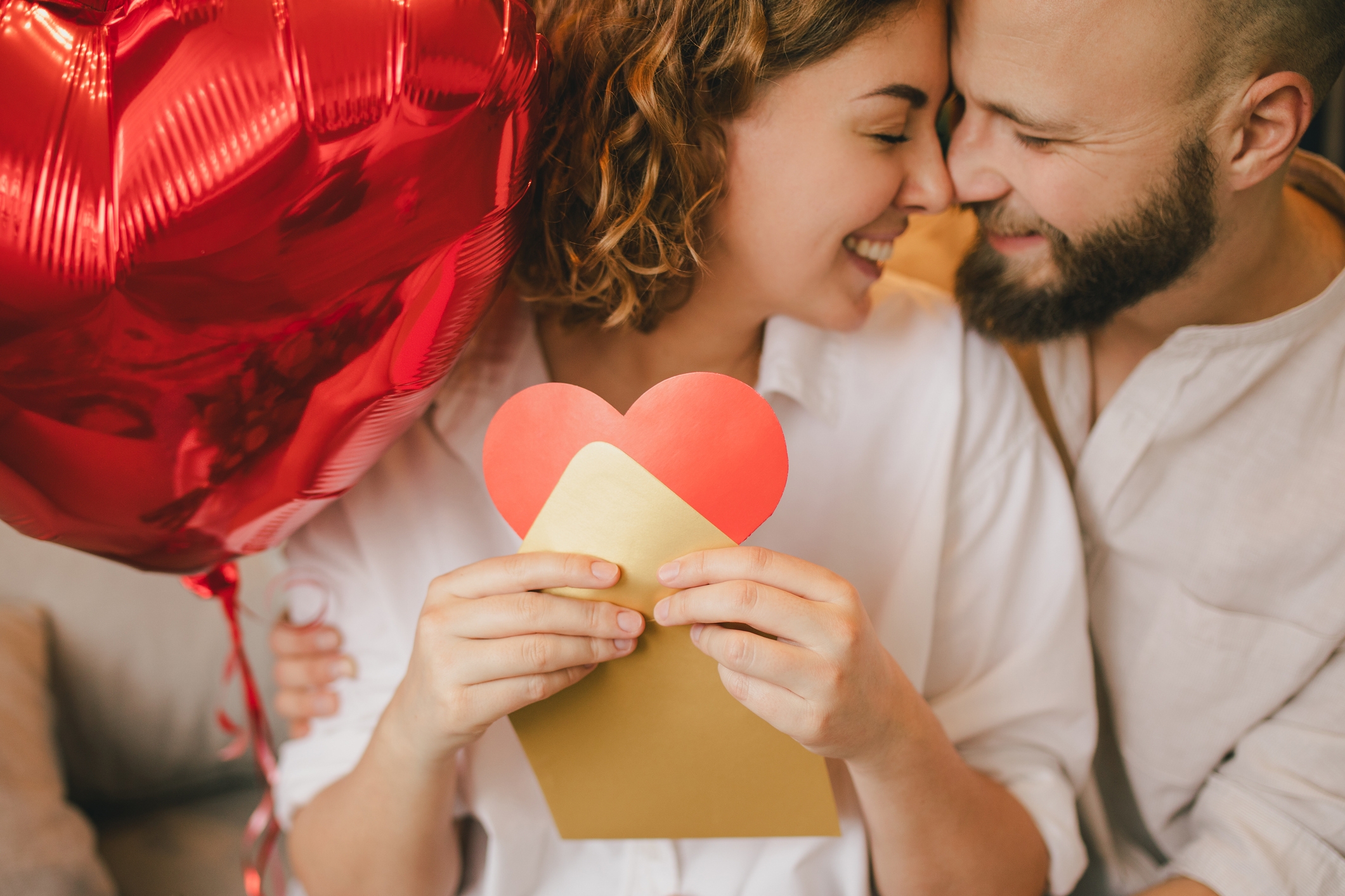 Couple smiles as they embrace, holding a red heart-shaped balloon and an envelope with a heart, conveying love and intimacy