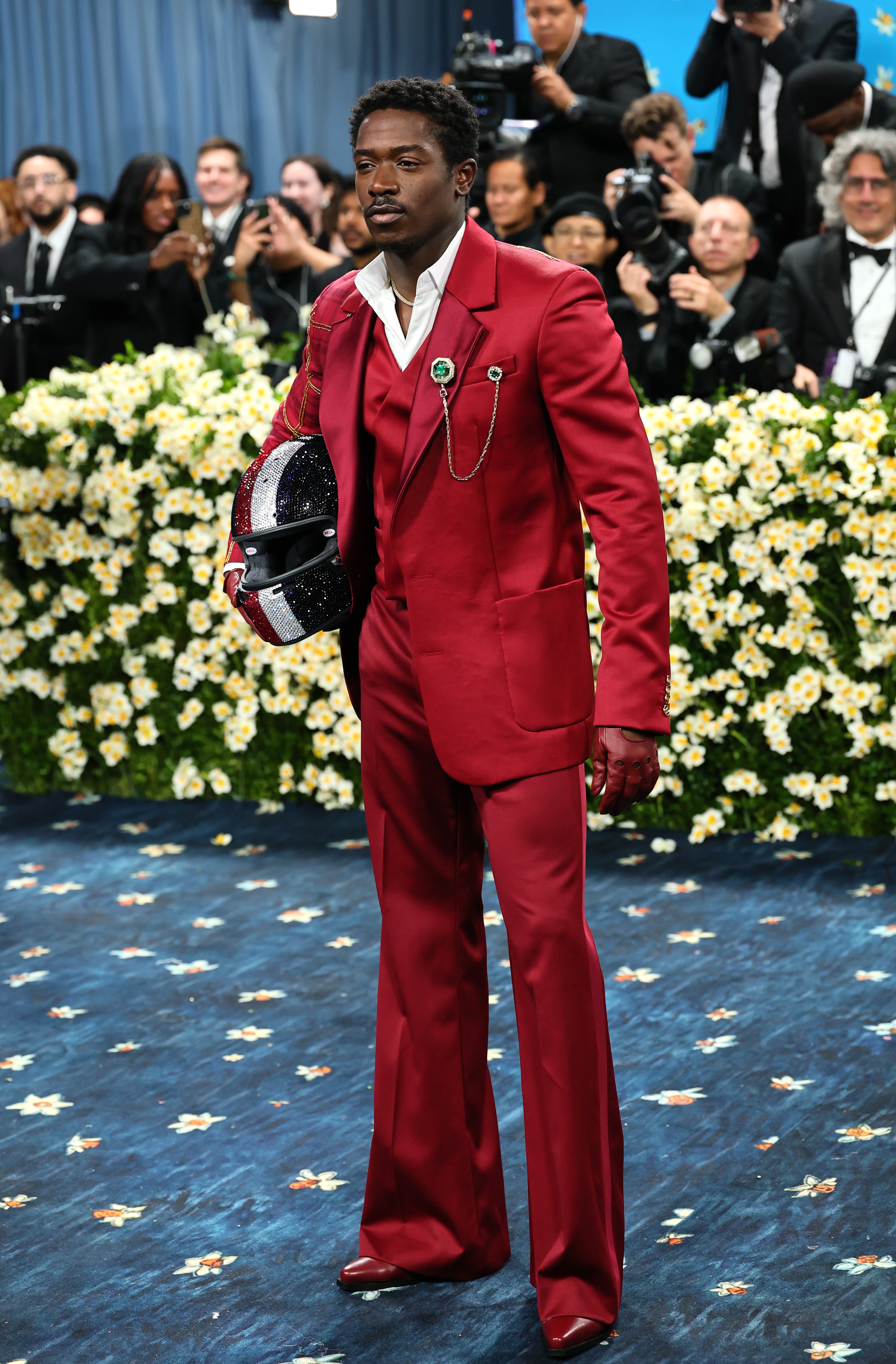 Person in a bold, tailored suit holds a sparkling helmet on a formal event carpet, surrounded by photographers and floral arrangements