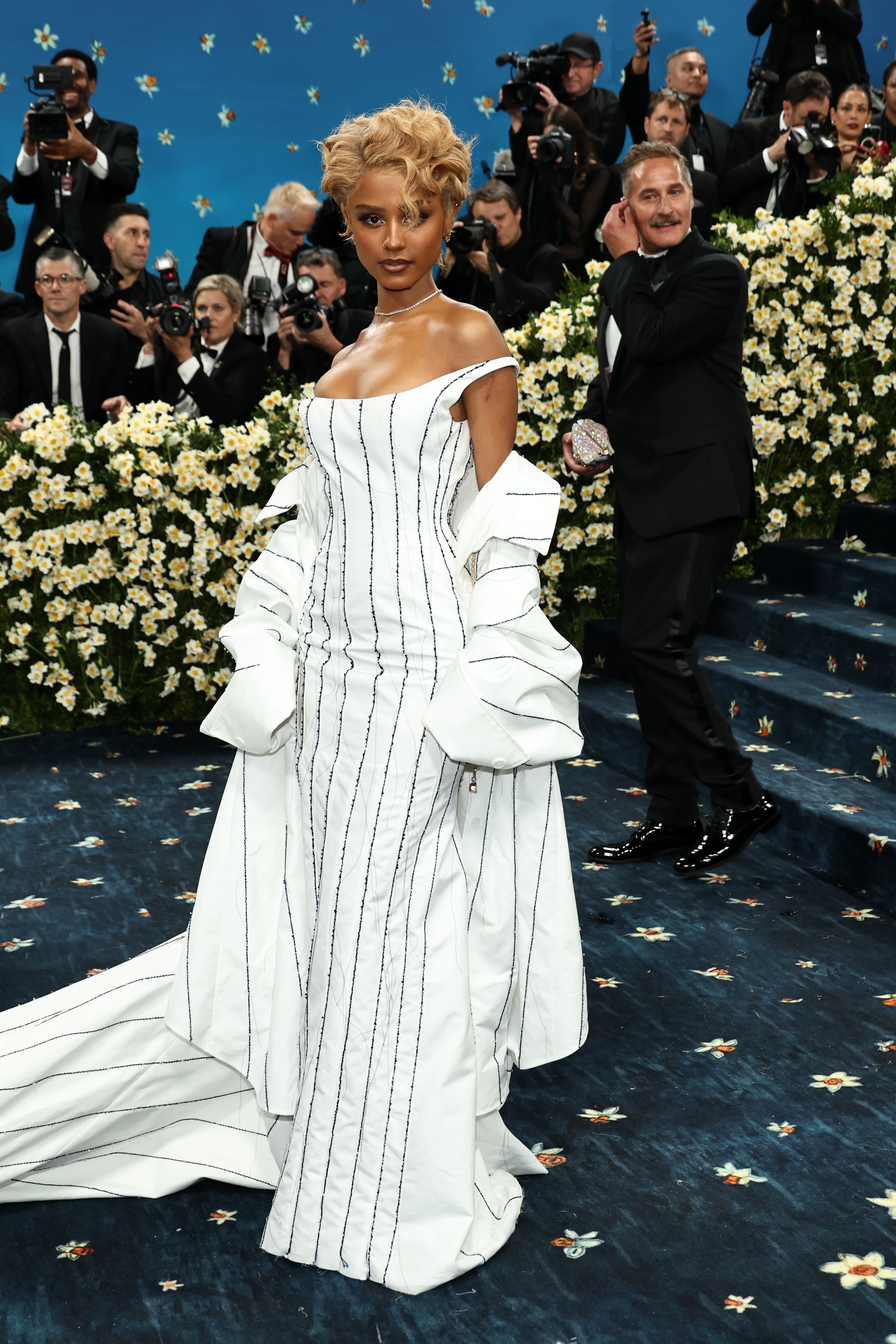 Person on a formal event carpet wearing an elegant, off-the-shoulder gown with vertical stripe details and long sleeves