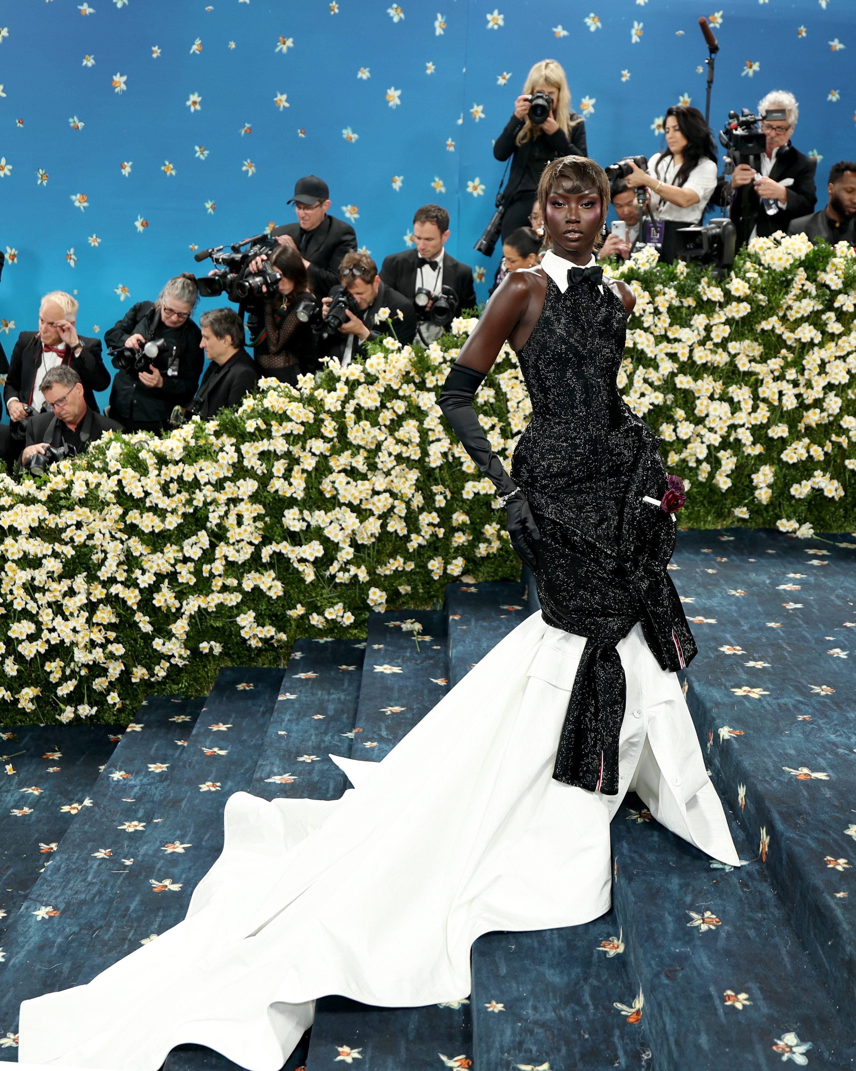 Person wearing an elegant, structured gown with a long white train poses on a floral-themed event carpet, surrounded by photographers