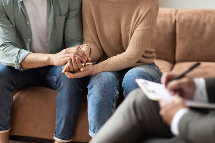 Couple holding hands in a counseling session, seated on a couch, with a therapist taking notes in the foreground