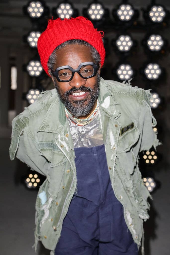 Person in eclectic, layered outfit with tattered jacket and beanie, smiling at camera