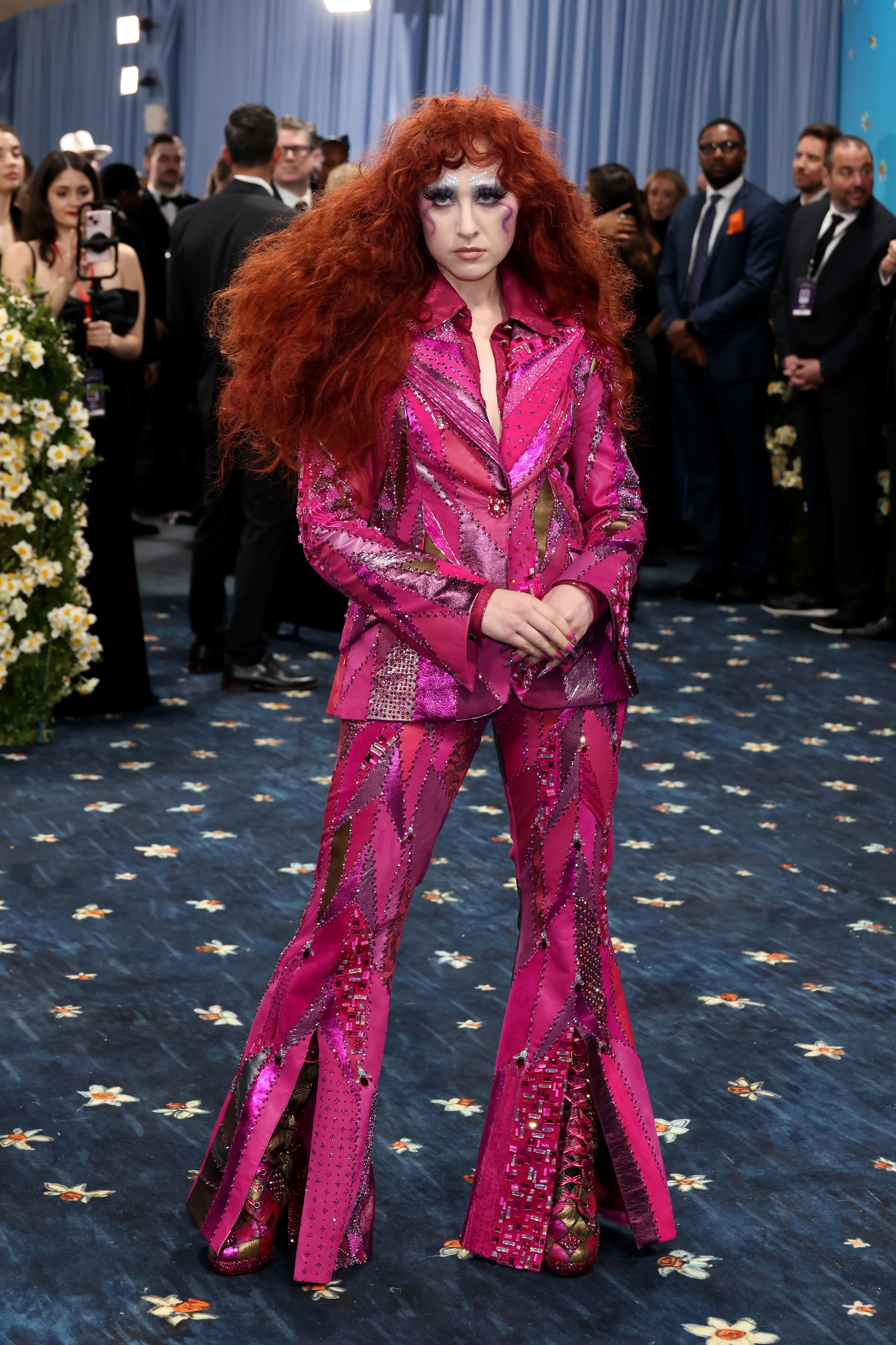 Person on blue carpet in bold, sequin-studded suit with flared pants and dramatic hairstyle, surrounded by attendees and floral decor