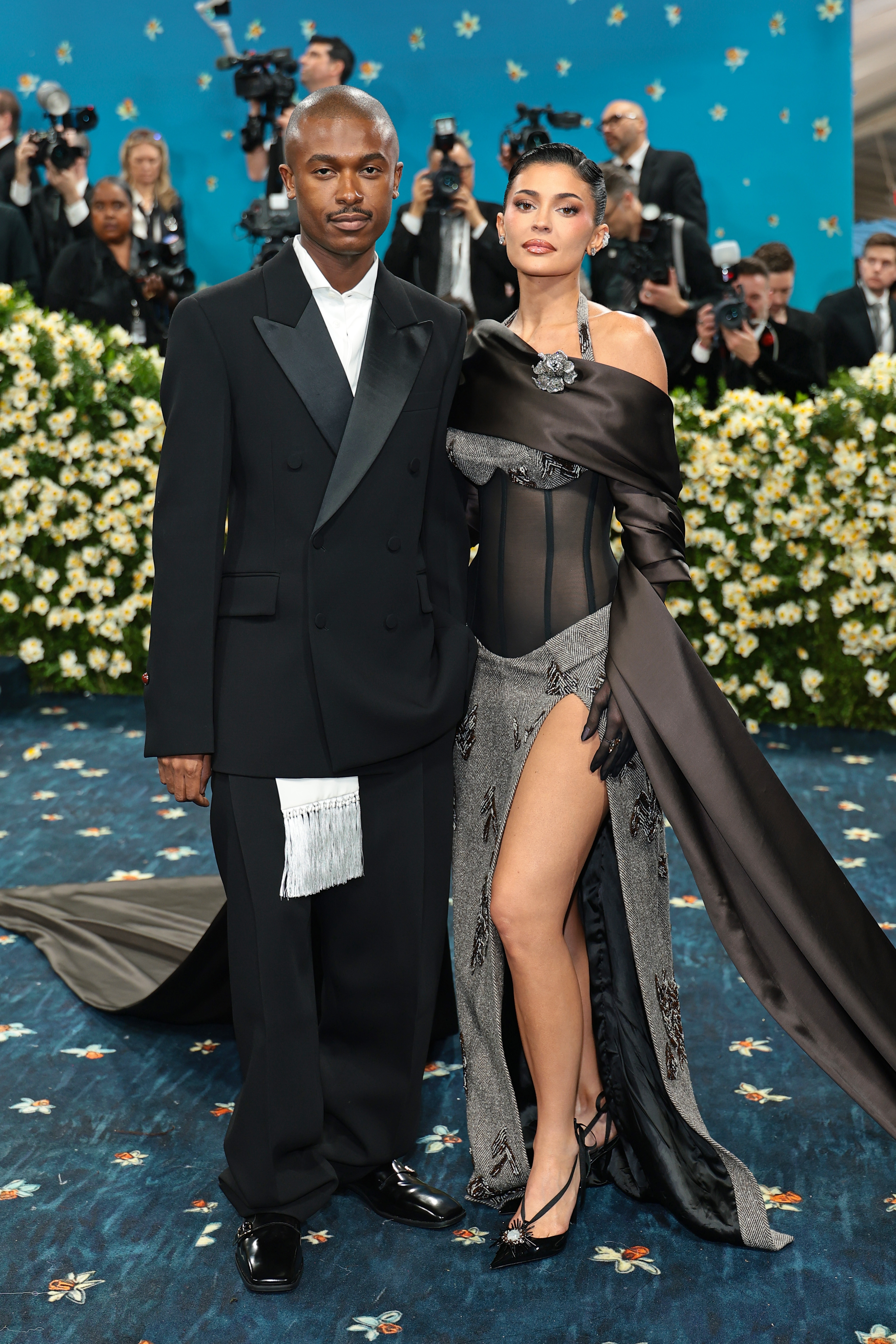 Man in a tailored suit and woman in a high-slit gown with sheer details on a formal event carpet