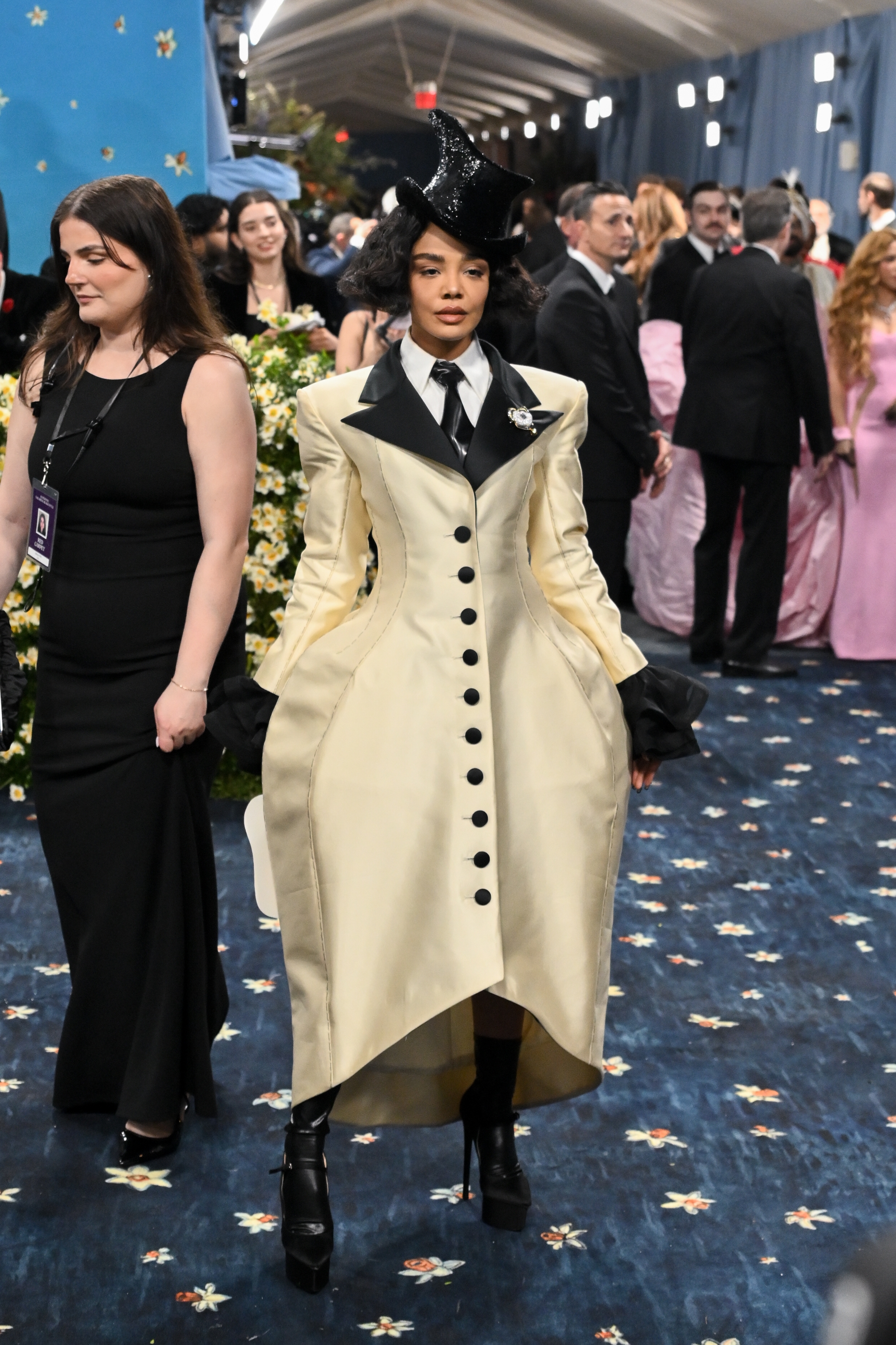 Celebrity in Victorian-inspired gown with high collar, exaggerated hips, top hat, and sleek boots on a formal event carpet