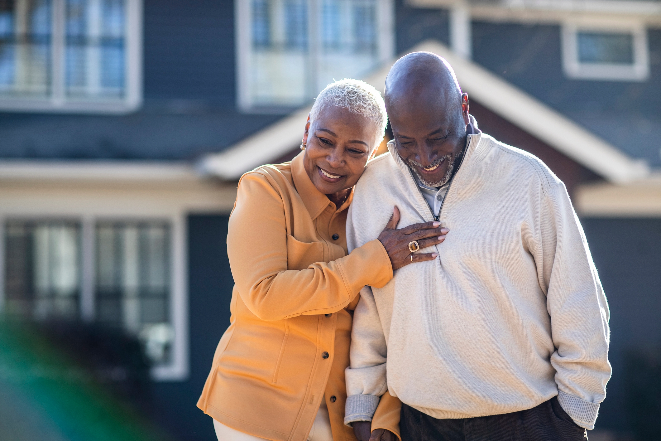 An older couple shares a joyful moment, smiling and leaning close in front of a house, symbolizing love and companionship
