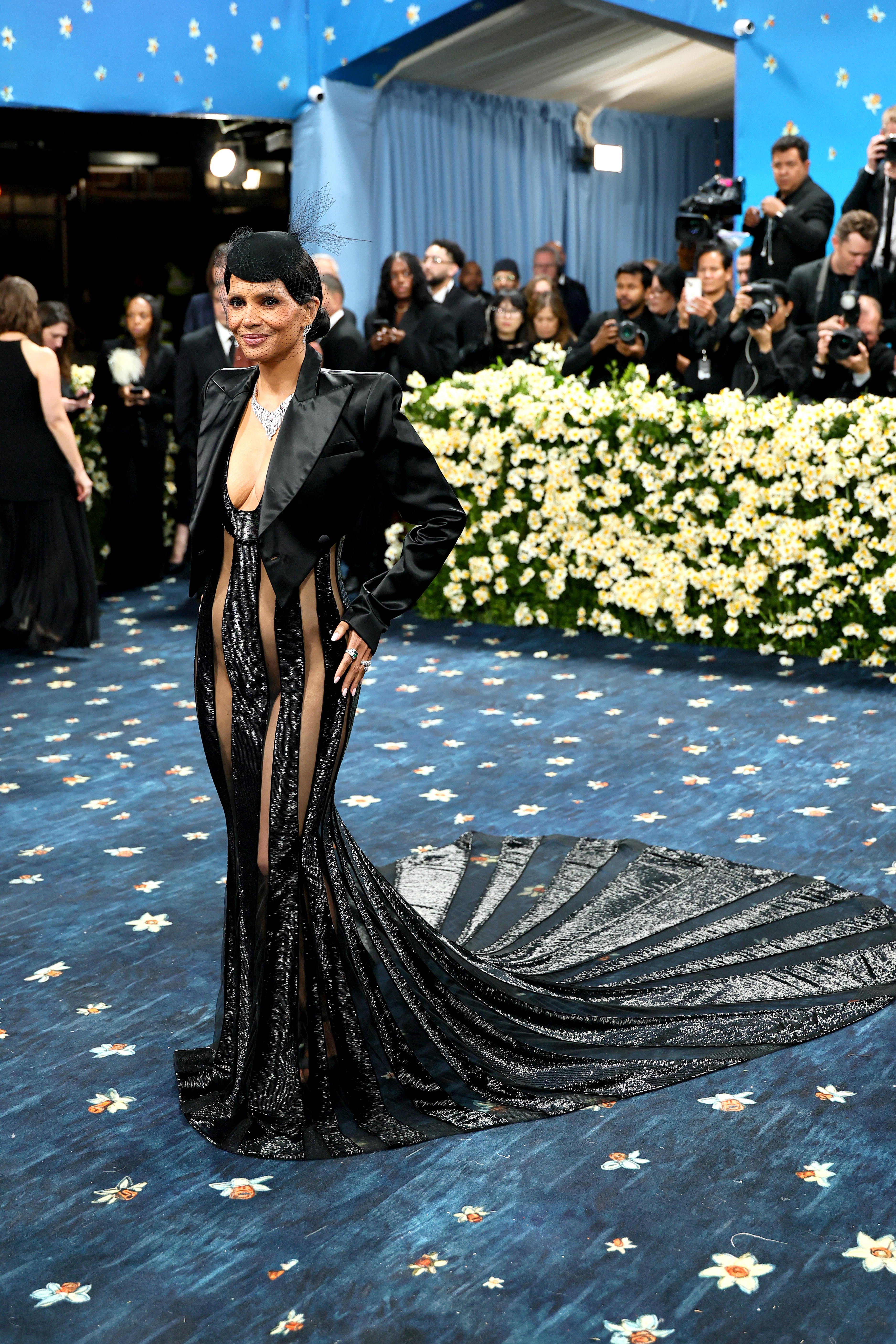 Celebrity in an elegant, form-fitting gown with sheer panels and a long train on a formal event carpet; people and photographers in the background