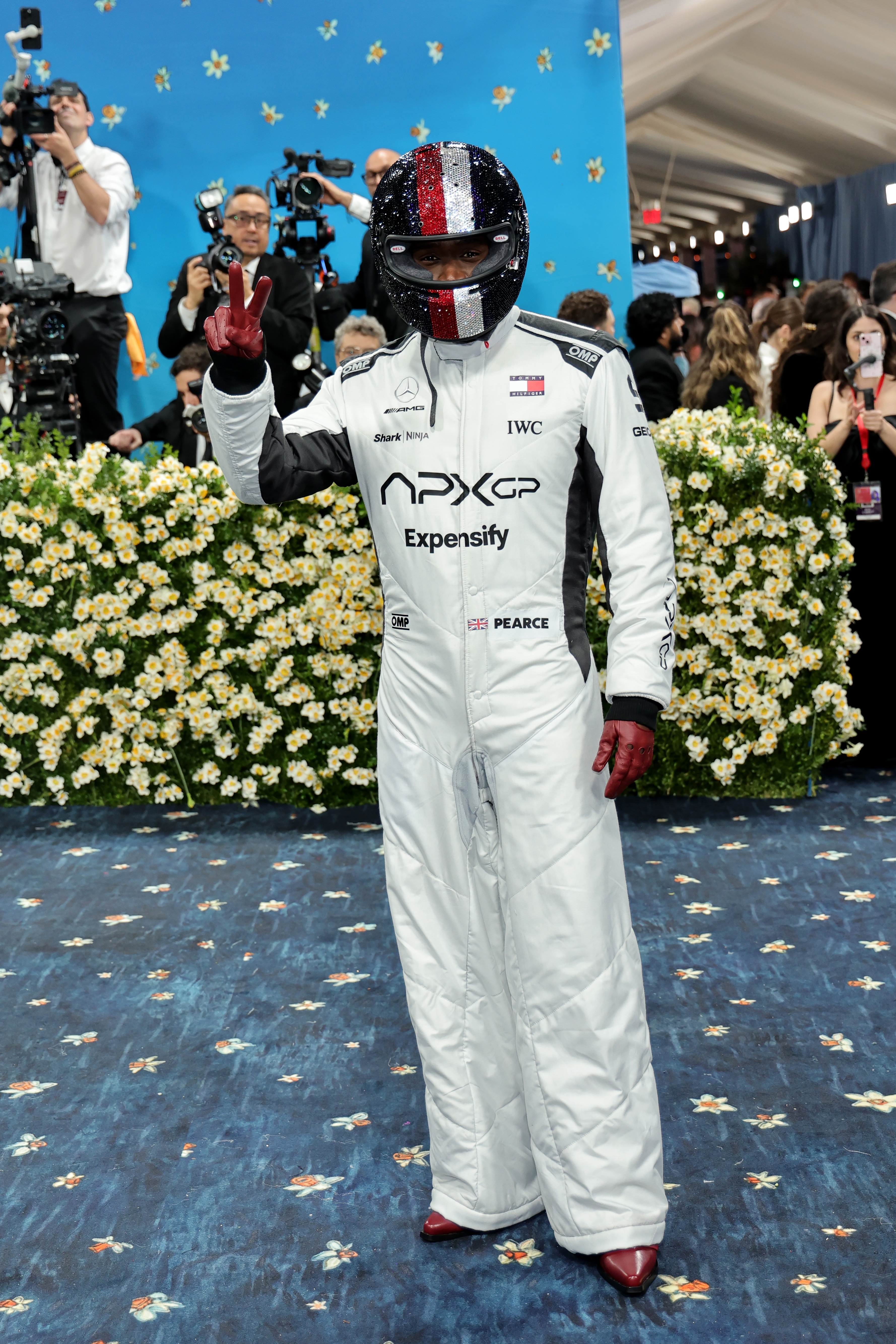 Person in full racing suit and helmet making a peace sign on an event carpet surrounded by photographers and floral decorations