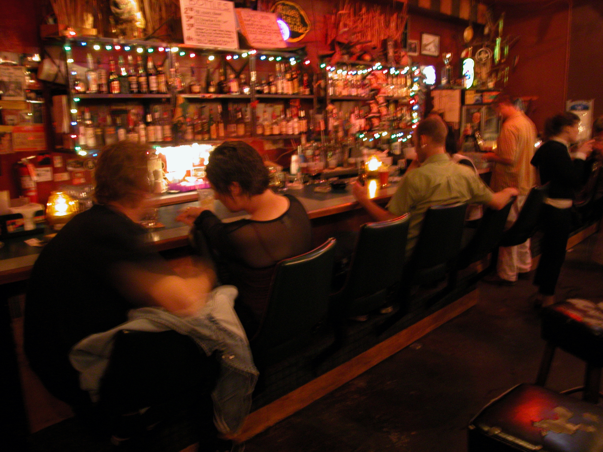 People sitting at a dimly lit bar with various bottles and decor in the background, engaged in conversations