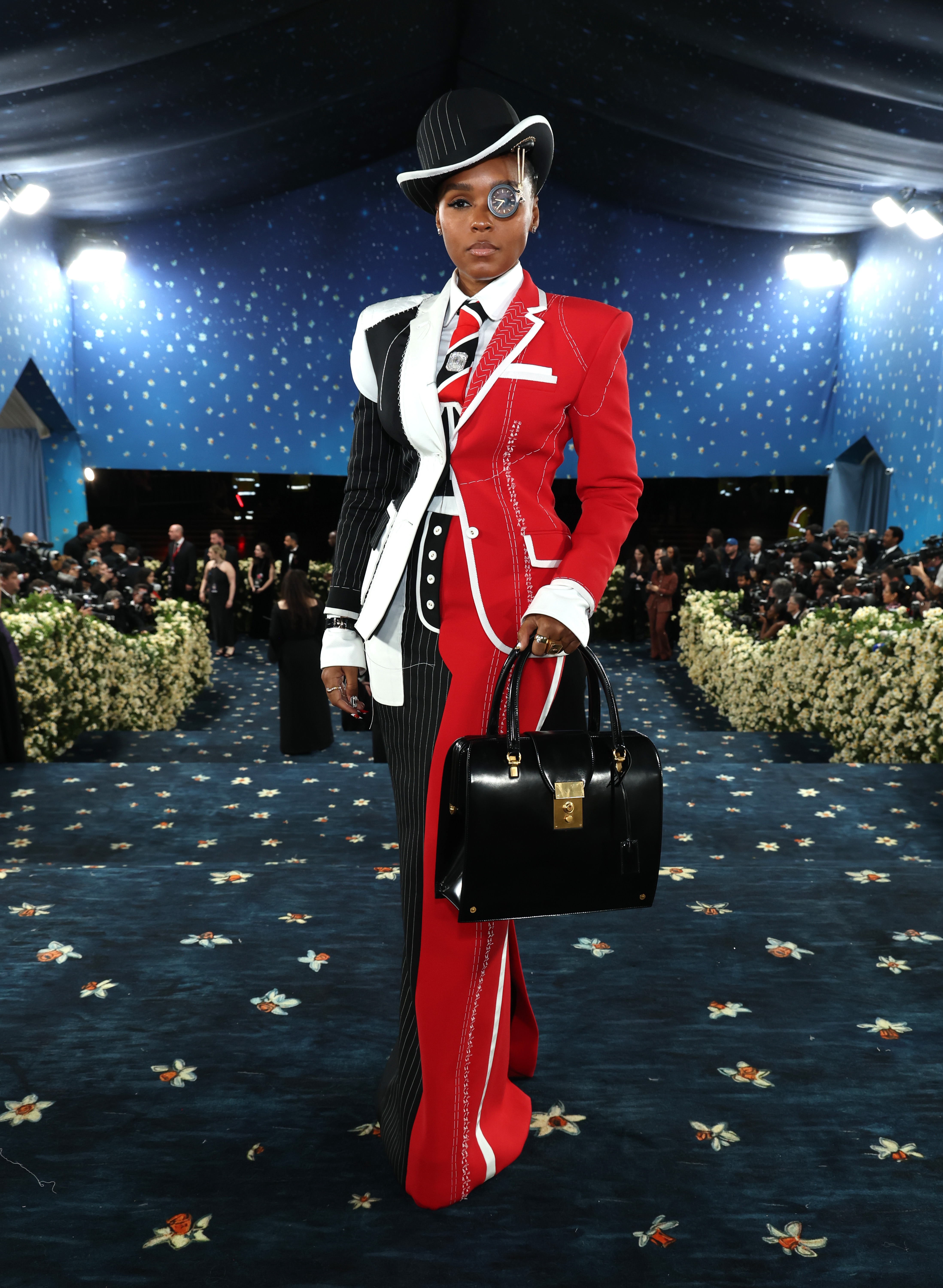 Person in a stylish, tailored outfit with pinstripes and color blocks, wearing a hat and monocle, on a formal event carpet