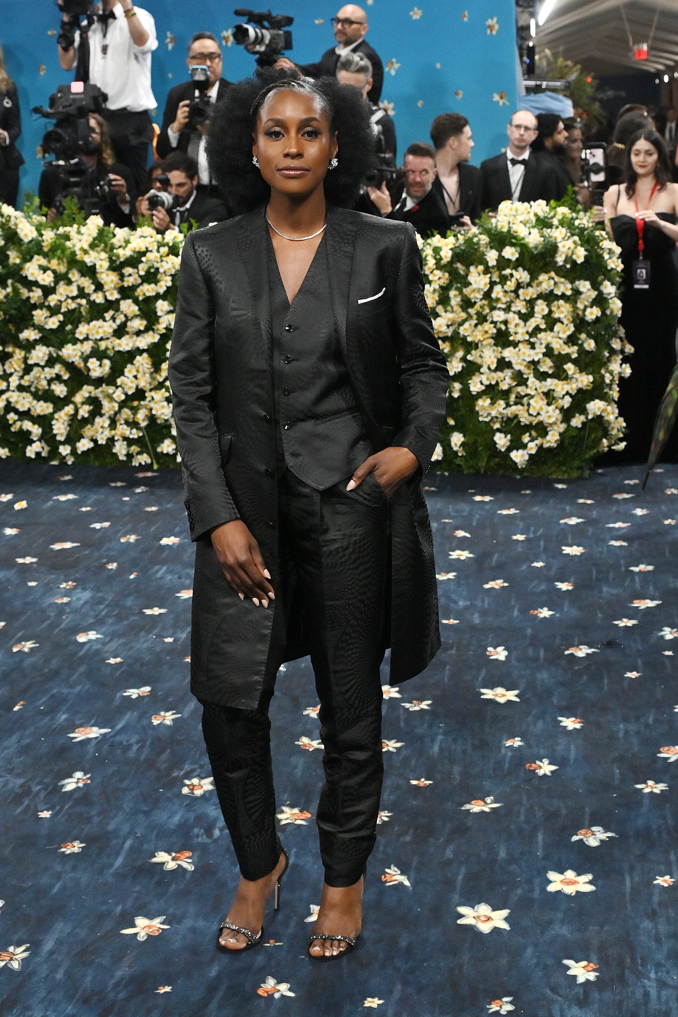 Person in a sleek, dark three-piece suit poses confidently on a floral-patterned carpet, surrounded by photographers at a celebrity event