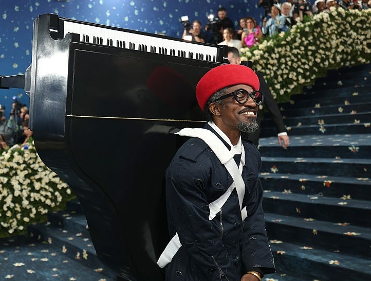 A person in a unique outfit resembling a piano, featuring a long coat and red beret, walks on a floral-decorated runway or event space