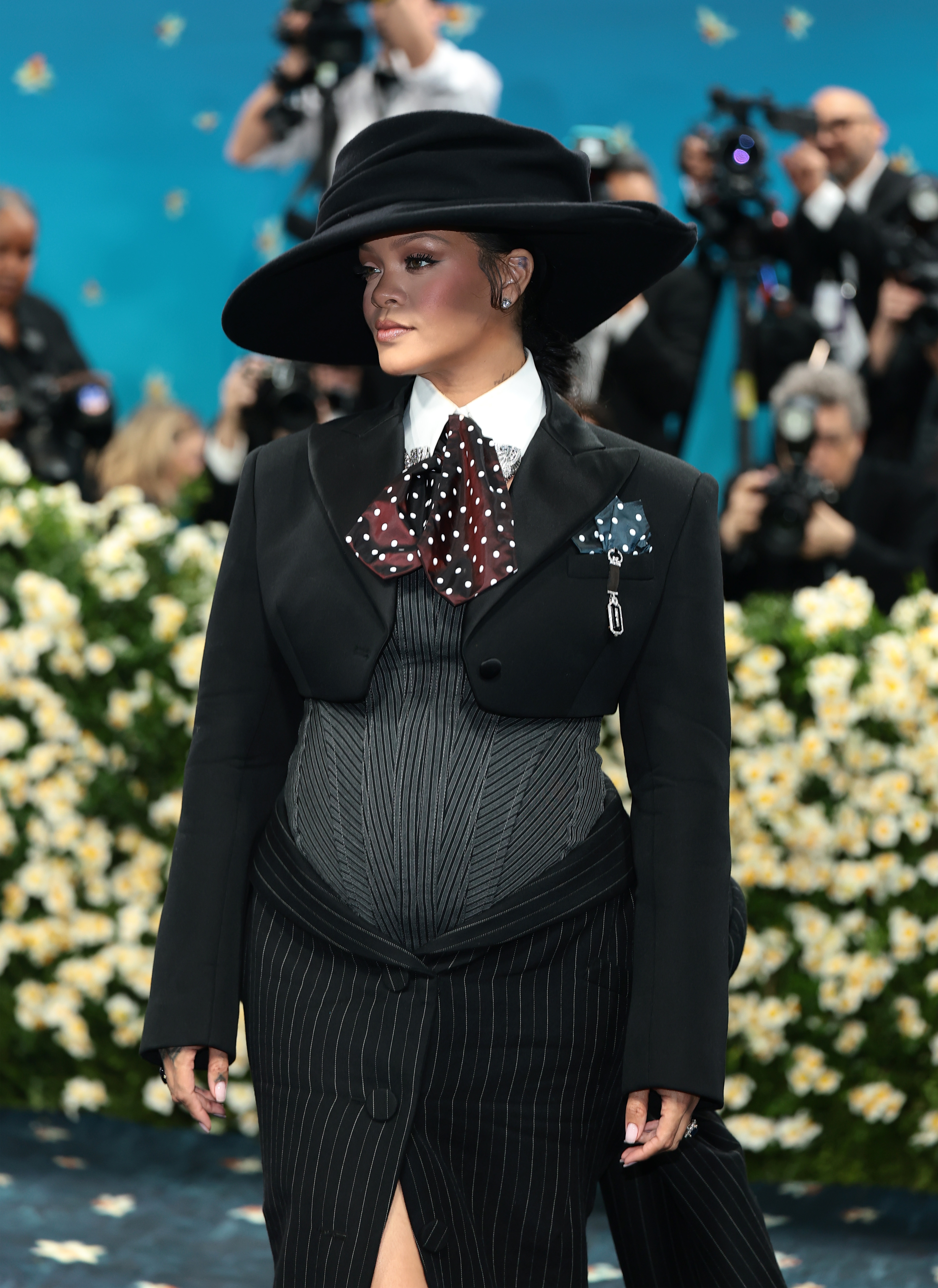 Person in vintage-inspired black suit and hat on a red carpet, with photographers in the background
