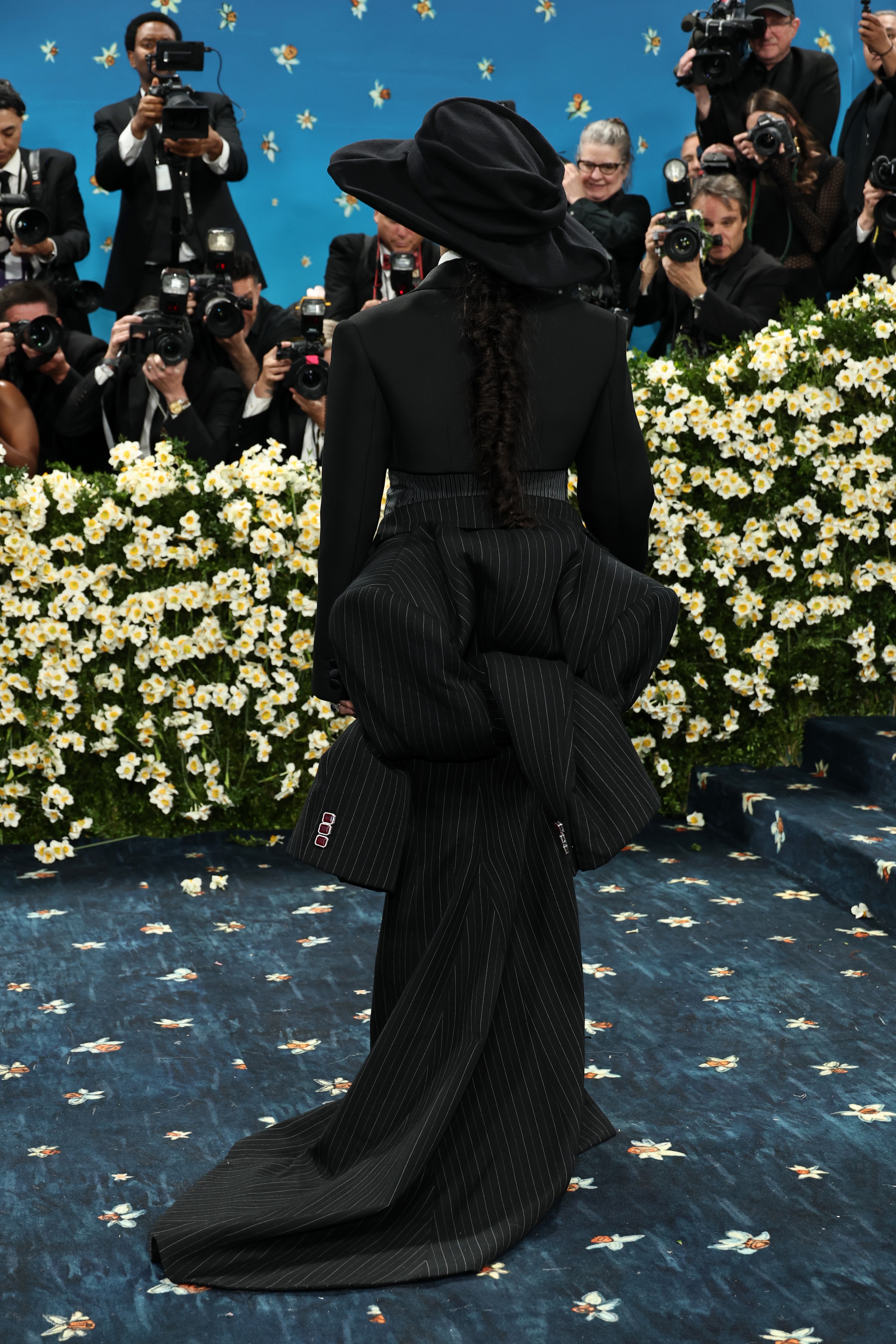 Person in an avant-garde pinstripe outfit with a large bow on the back, posing at an event, surrounded by photographers and flowers