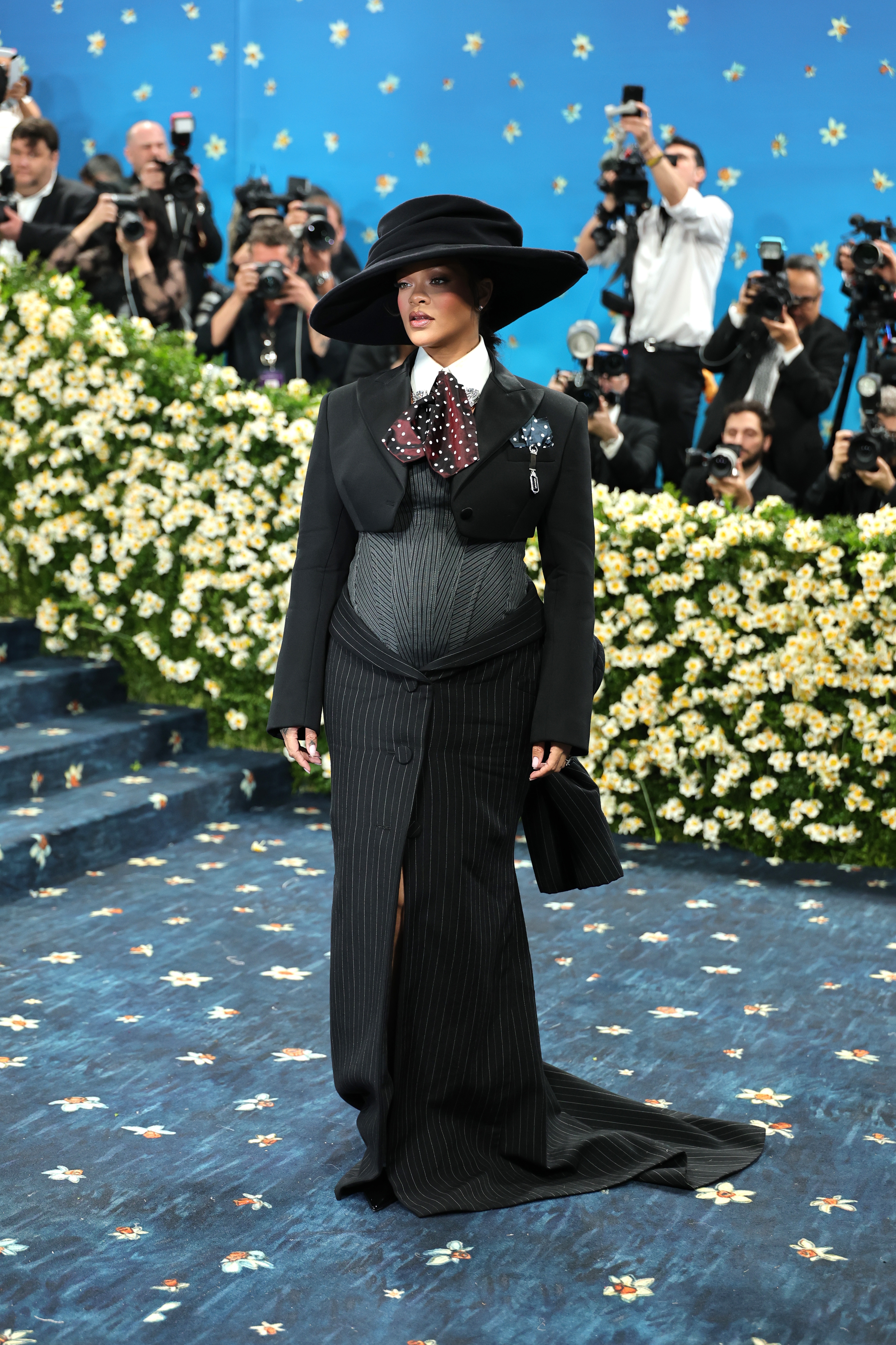 Person in a tailored suit-dress with pinstripes and a large hat on a floral-adorned carpet, surrounded by photographers