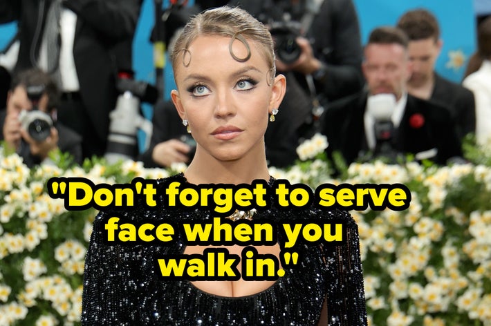 Person in a sequined gown with shoulder detail and a circular neckline on the red carpet, surrounded by photographers and floral decor