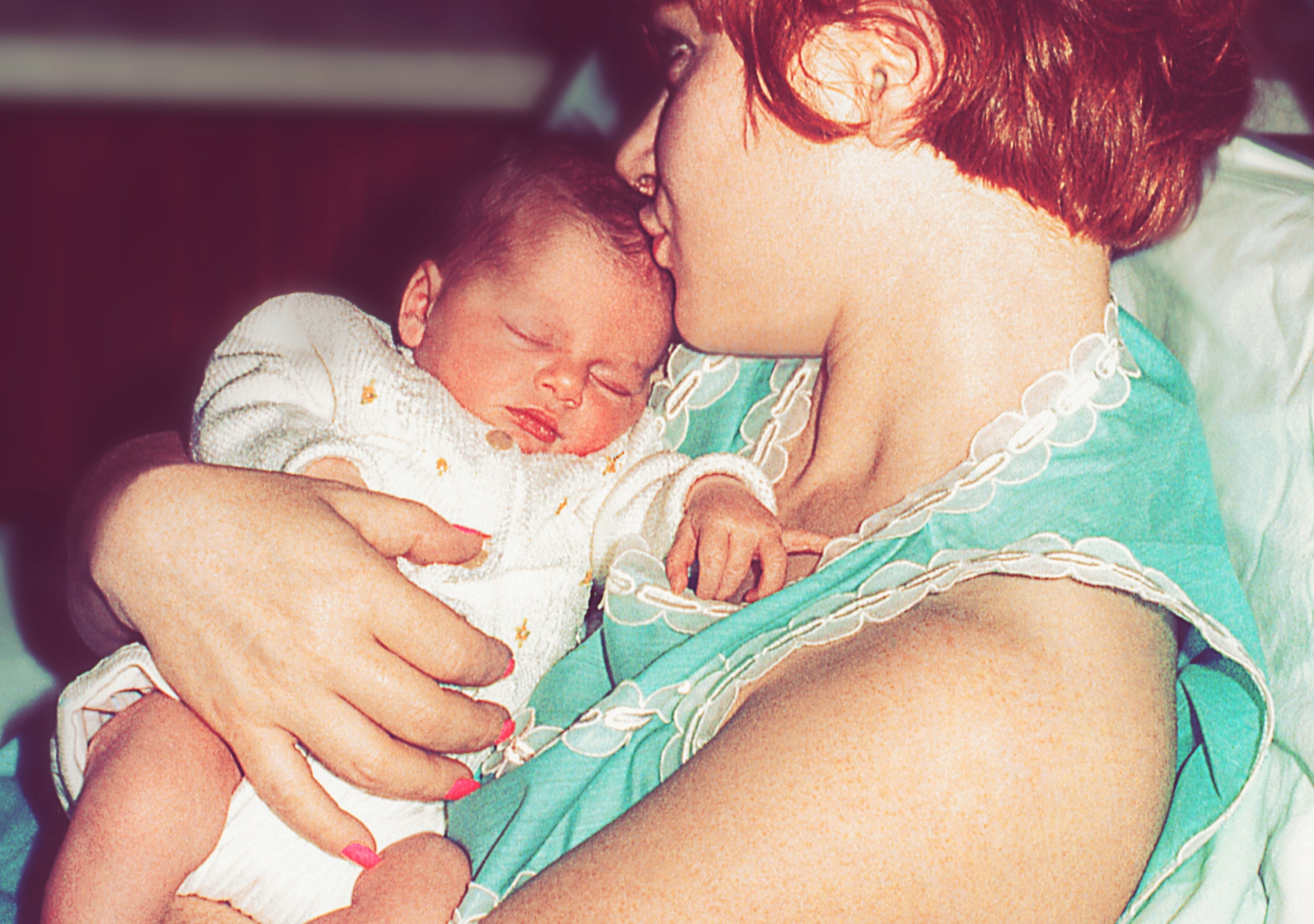 A woman with short hair cradles a sleeping baby in her arms, both wearing hospital tags, suggesting a post-birth setting