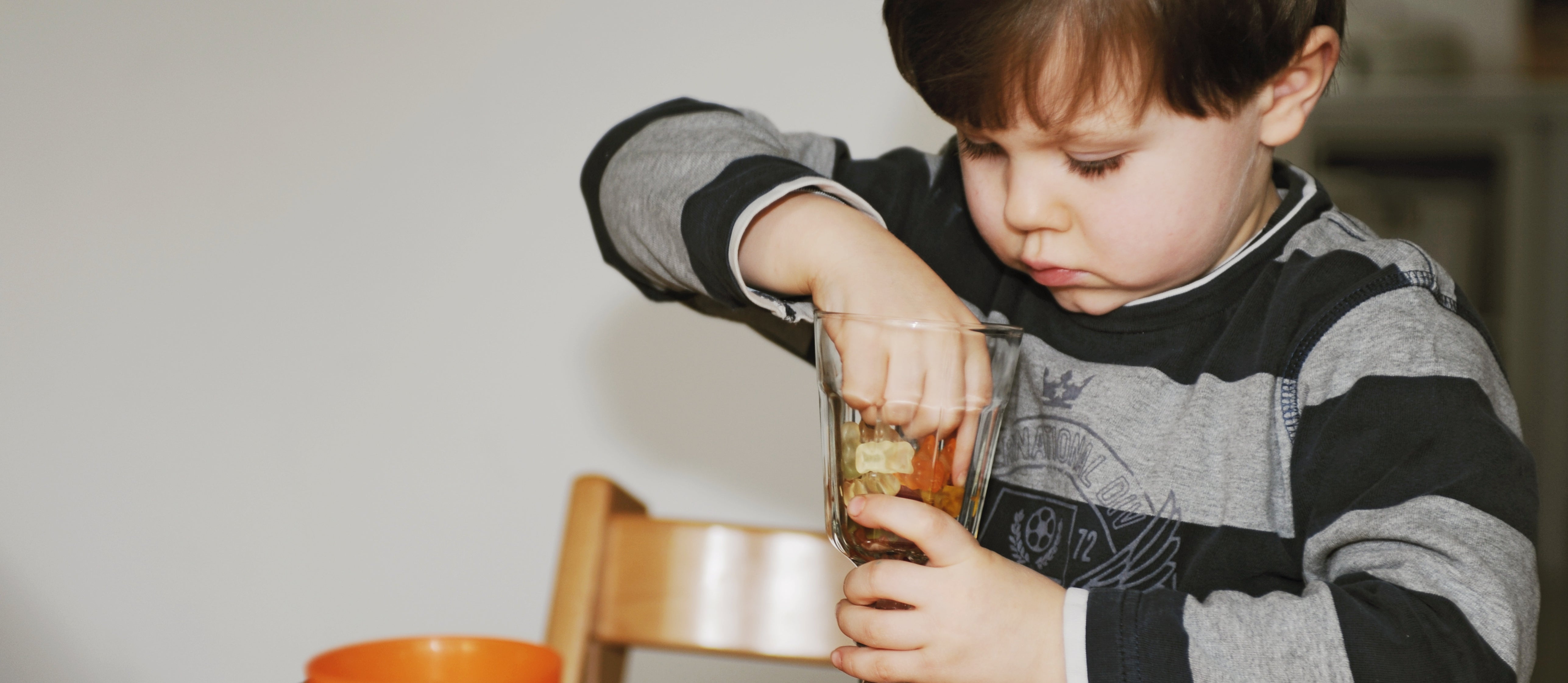 Child sitting at a table, reaching into a glass with candies inside, looking focused. An orange cup and more candies are on the table