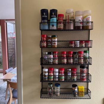 Wall-mounted spice rack with four shelves, holding various seasoning bottles and containers, next to a dining area